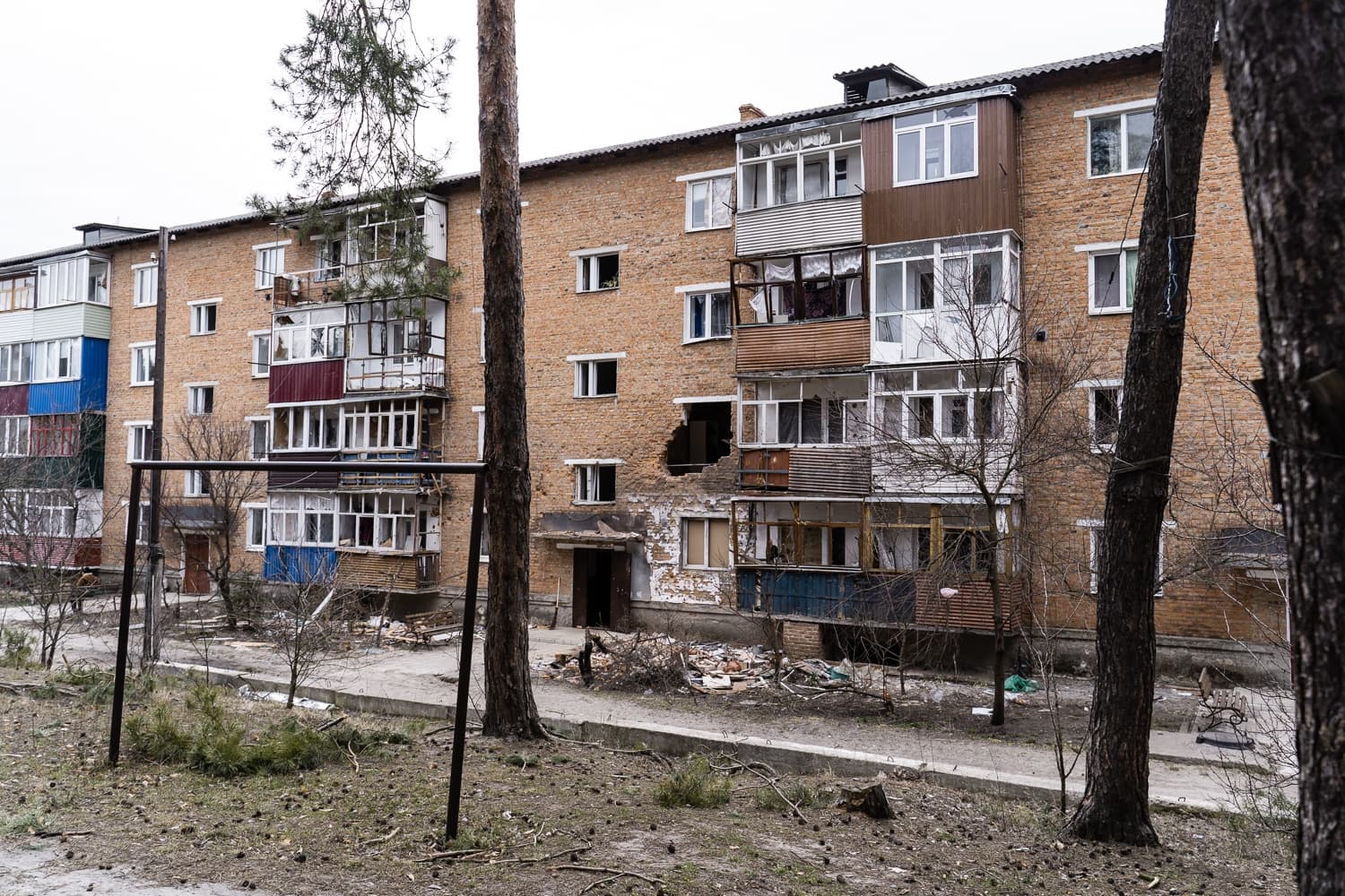 Поврежденный обстрелом кирпичный жилой дом с проломом в стене, виден сквозь сосны.