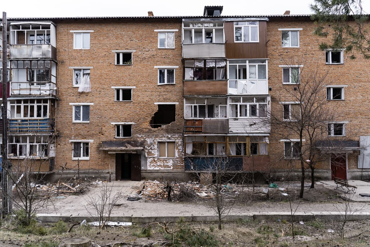 Разрушенный обстрелом кирпичный многоквартирный дом с выбитыми окнами, поврежденными балконами и большой пробоиной в стене.