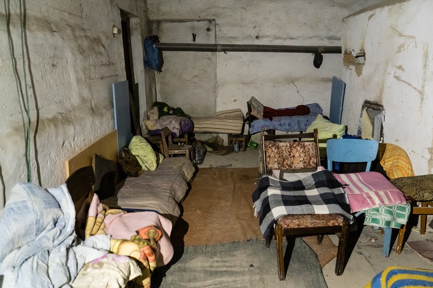 Тесное, грязное подвальное помещение, используемое как импровизированное убежище, с кроватями, матрасами и стульями, покрытыми одеялами.