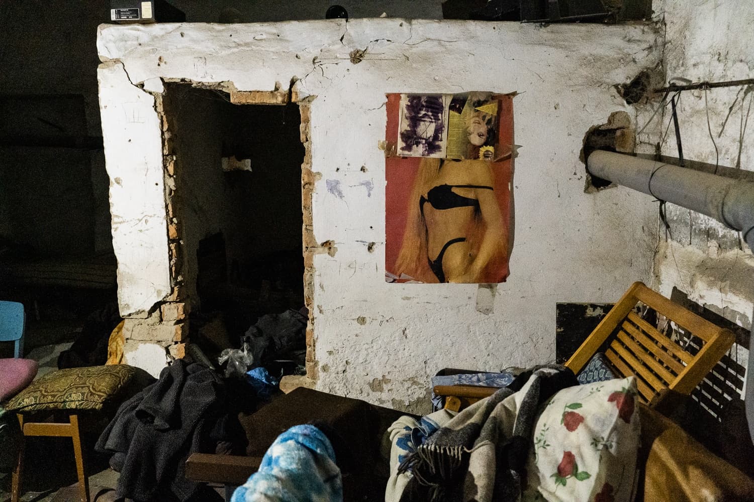 Разрушенное помещение с осыпающейся белой стеной и открытым дверным проемом. На стене плакат с женщиной в черном бикини. Пол завален одеялами и мебелью.