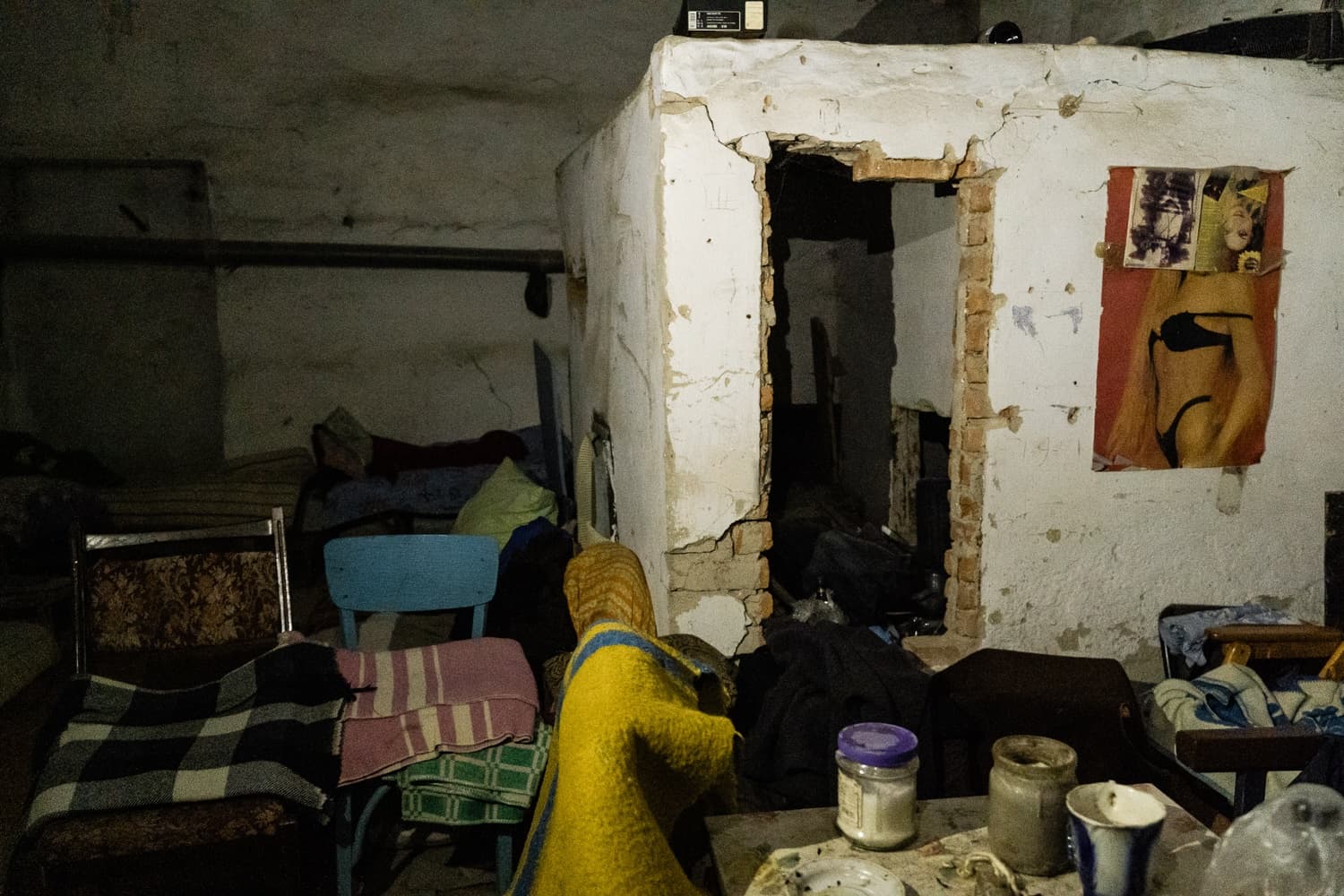 Темное, захламленное убежище: матрасы, одеяла, стулья, обвалившийся дверной проем с кирпичом, плакат на стене.