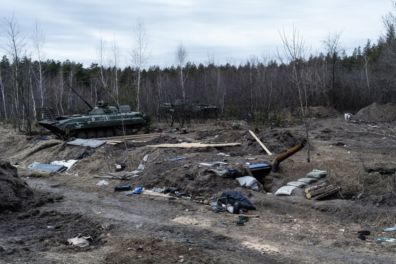 Военная техника БМП-1 стоит на фоне леса, рядом с разрушенными блиндажами и окопами.
