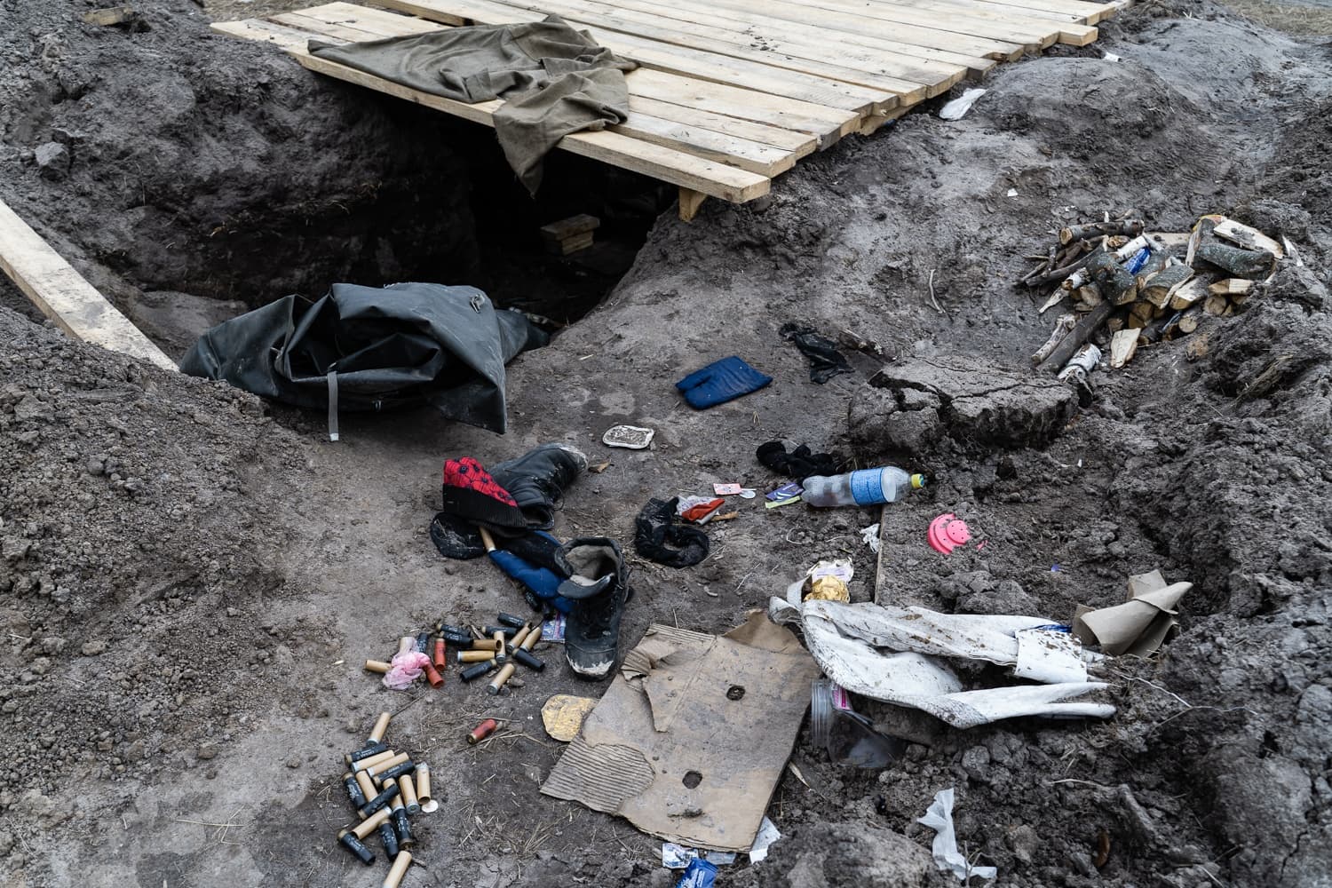 Окоп или землянка с деревянным настилом, вокруг разбросаны гильзы, обувь, одежда, бутылка и мусор.