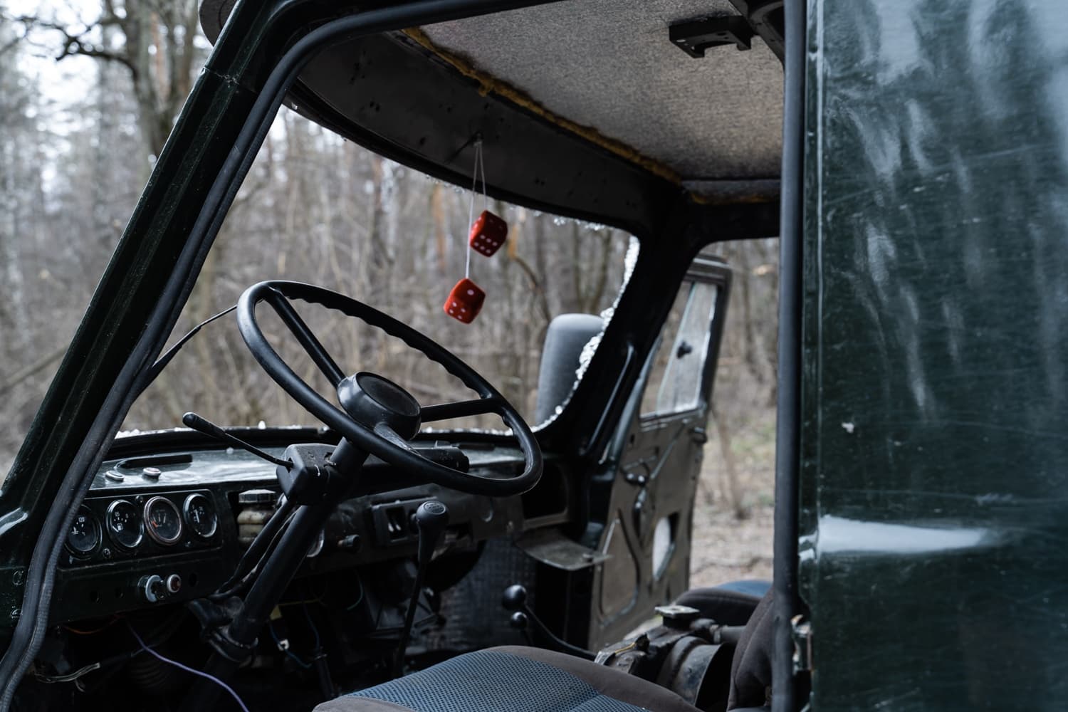 Интерьер кабины старого грузовика (возможно, УАЗ) с приборной панелью, рулем и красными игральными костями, свисающими с потолка; на заднем плане лес.