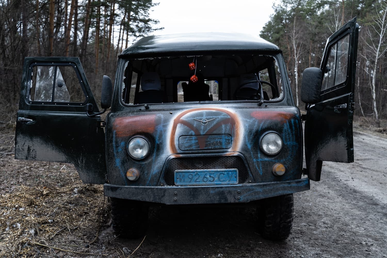 Разбитый УАЗ-452 с граффити на капоте, открытыми дверями и подвешенными игральными костями, стоит на лесной дороге. Номер "М 92 65 СУ".