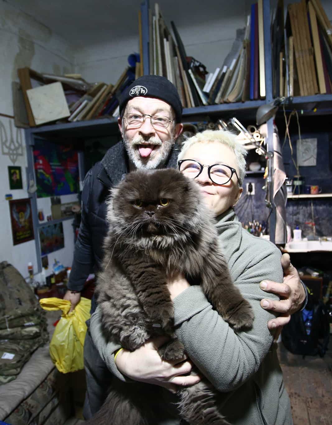 Женщина держит большого пушистого кота, мужчина стоит за ней, оба улыбаются, на фоне полки с вещами.