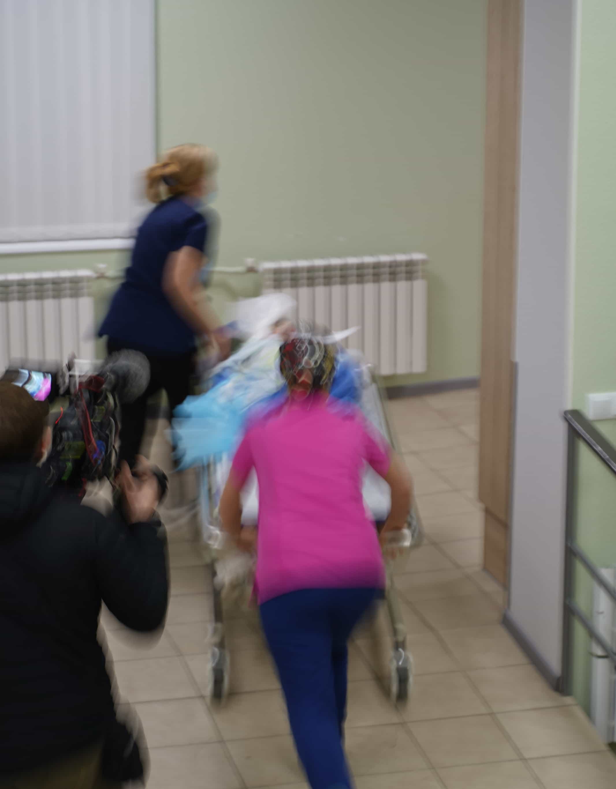 Размытое изображение медицинских работников, толкающих каталку в больничном коридоре.