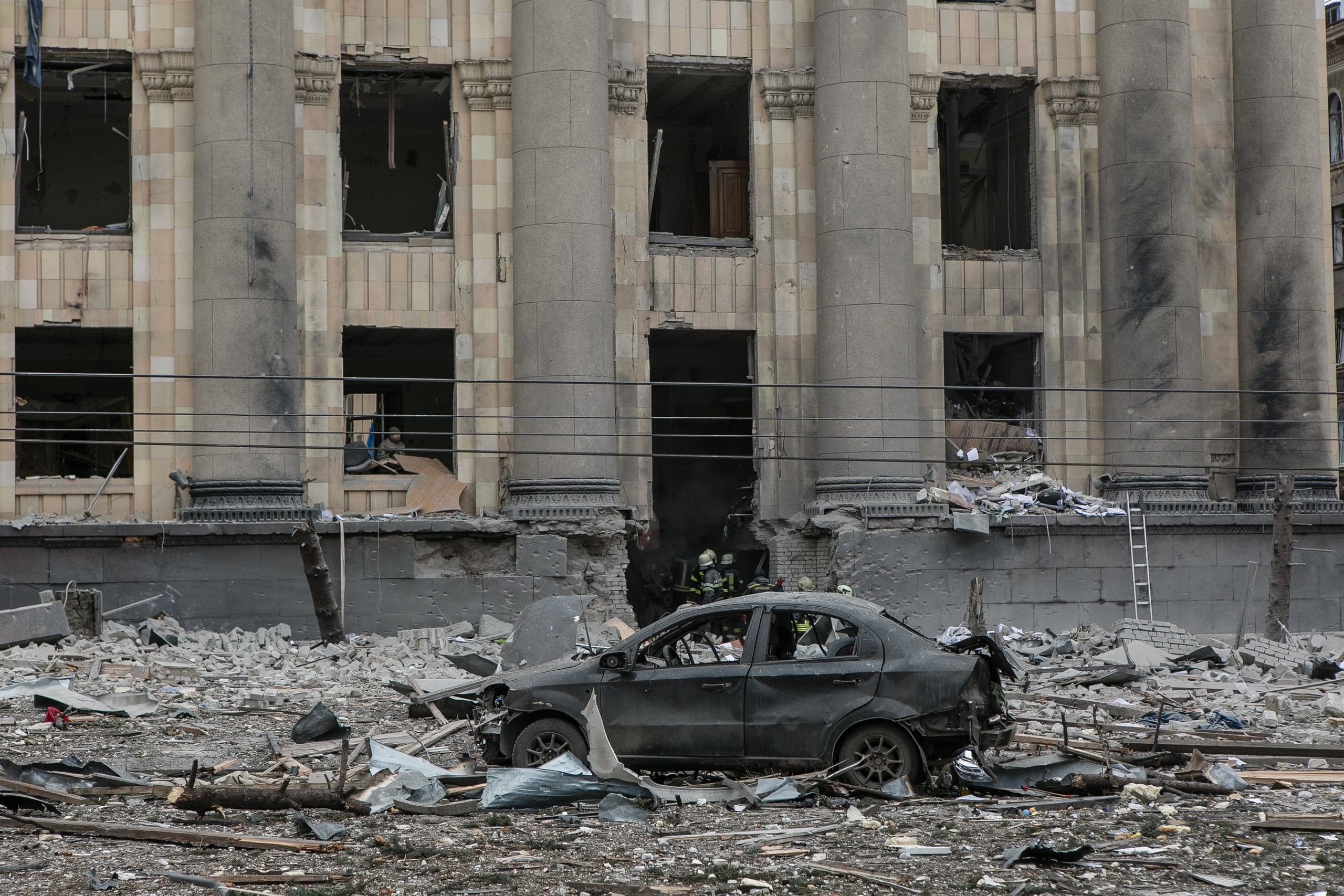 Разбитый автомобиль и обломки на фоне сильно поврежденного здания с колоннами. Видны спасатели.