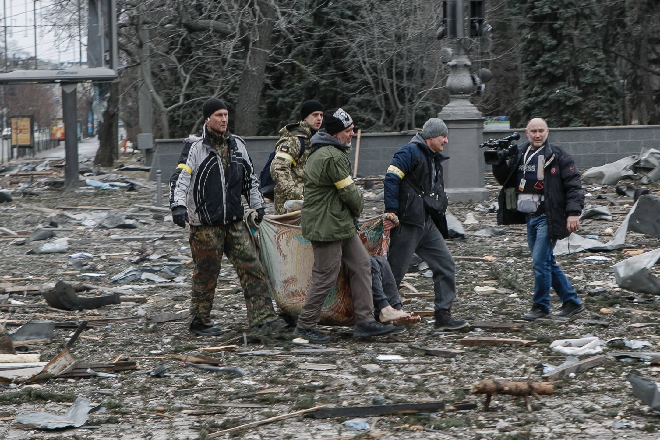 Четверо мужчин, некоторые с желтыми повязками, несут человека на ковре через завалы на улице. Справа оператор снимает их, на его бейдже надпись «PRESS».