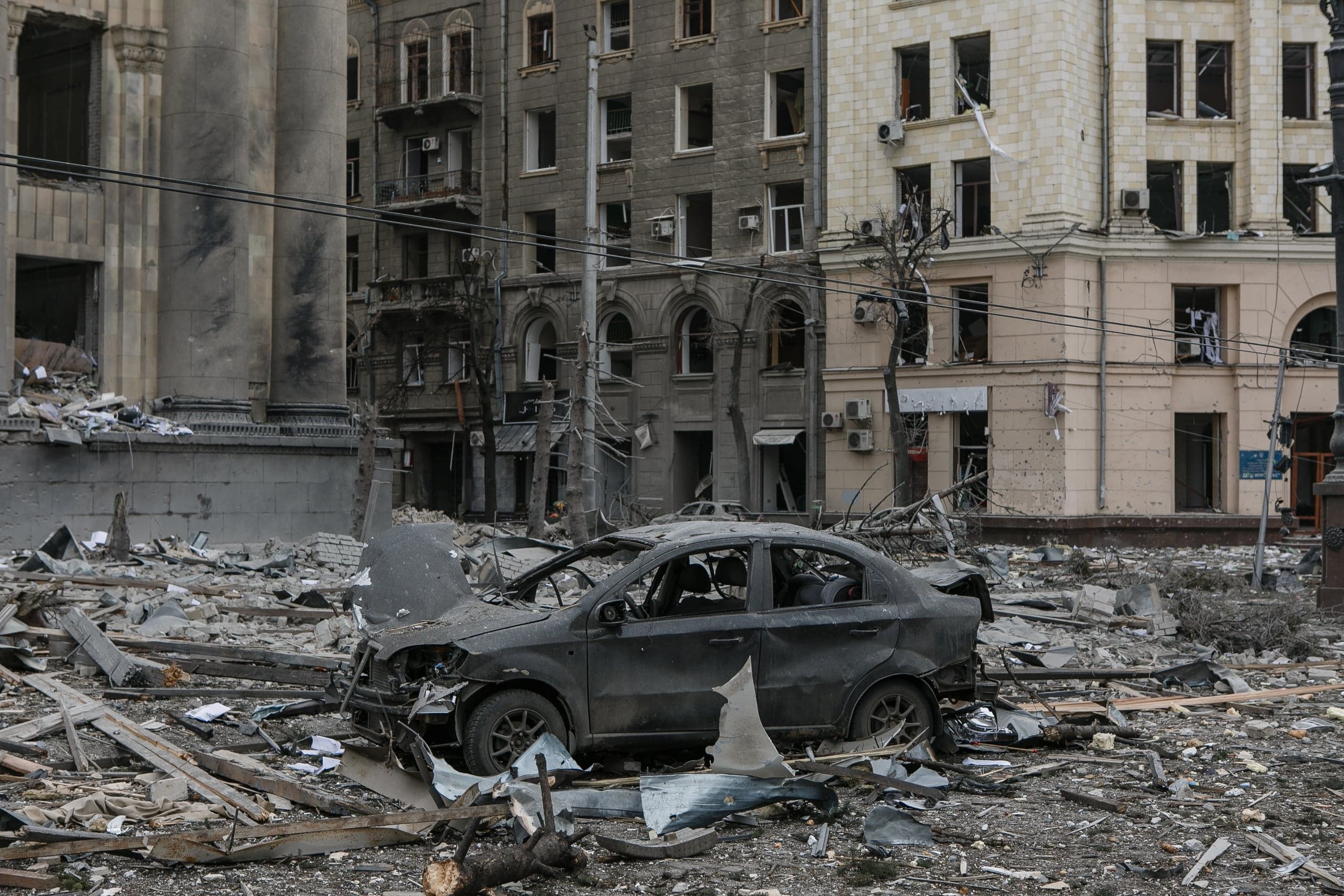 Улица, заваленная обломками, со сгоревшим автомобилем на фоне разрушенных зданий.