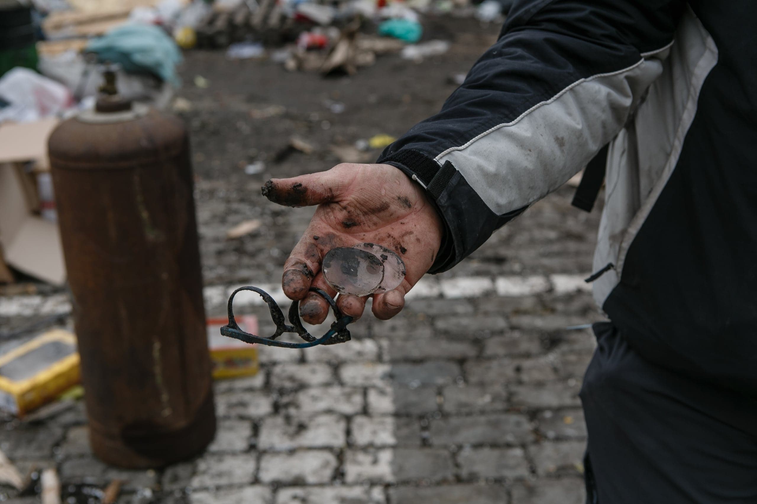 Грязная рука держит оплавленные и разбитые очки, на фоне разрухи, булыжников и ржавого газового баллона.