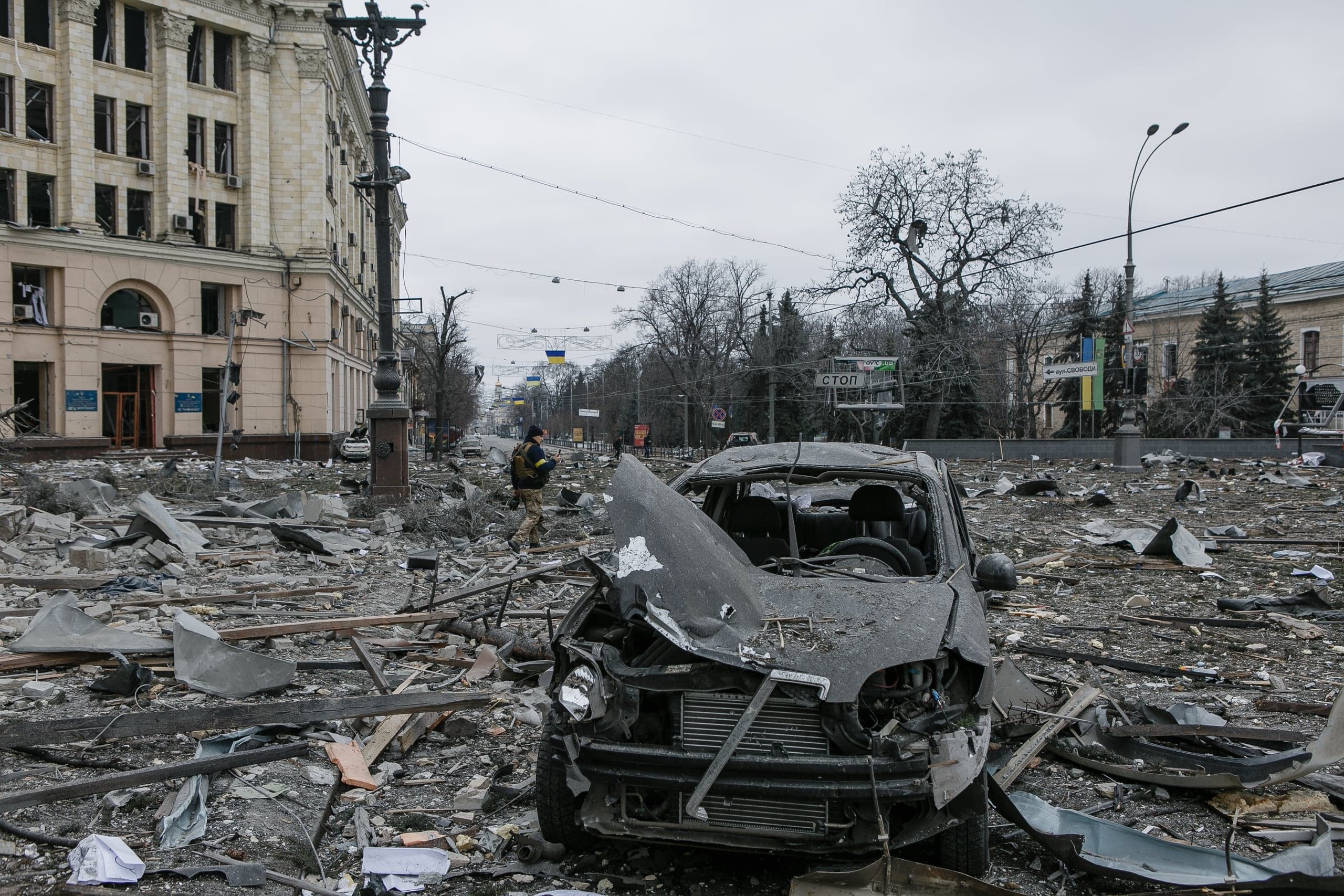 Разрушенный автомобиль посреди заваленной обломками площади в Харькове; на заднем плане виднеется поврежденное здание и военнослужащий. На дорожном знаке надпись: "СТОП".