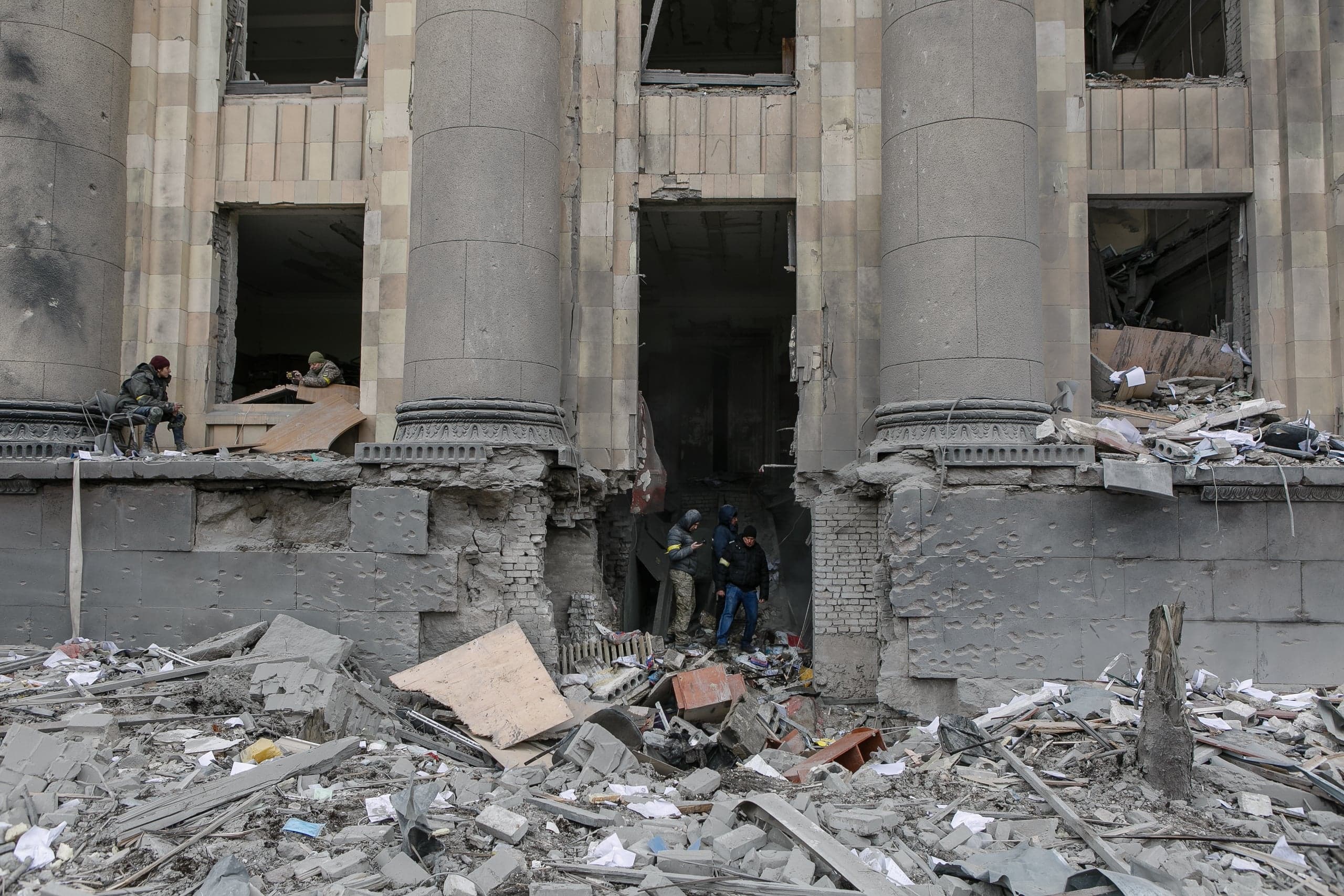 Фасад разрушенного здания с колоннами, пробоиной в стене и огромными завалами. Несколько человек видны в оконных и дверных проемах.