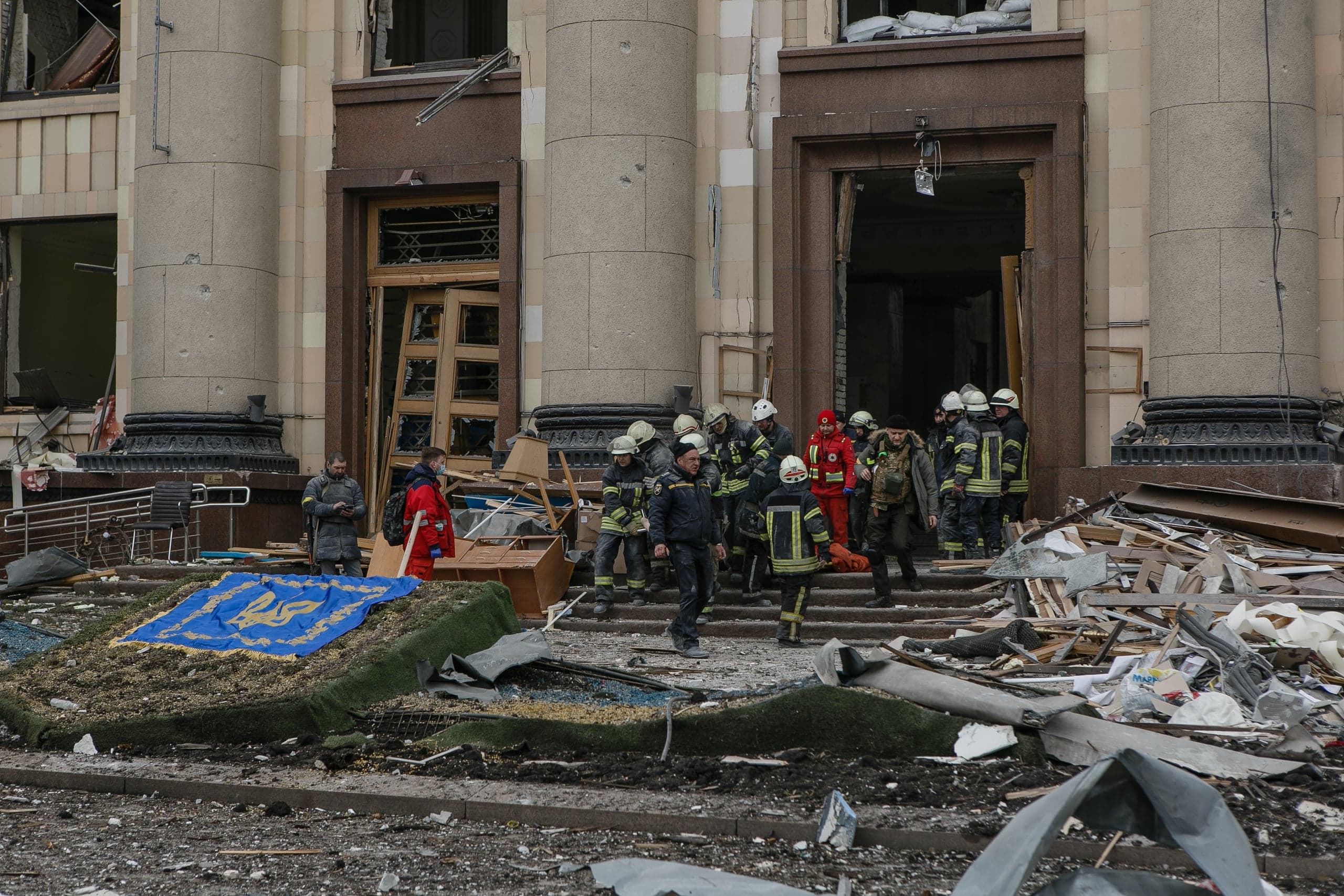 Последствия обстрела: группа спасателей у разрушенного входа в здание с колоннами. Обломки на ступенях и на земле. В центре синий ковер с украинским тризубом.