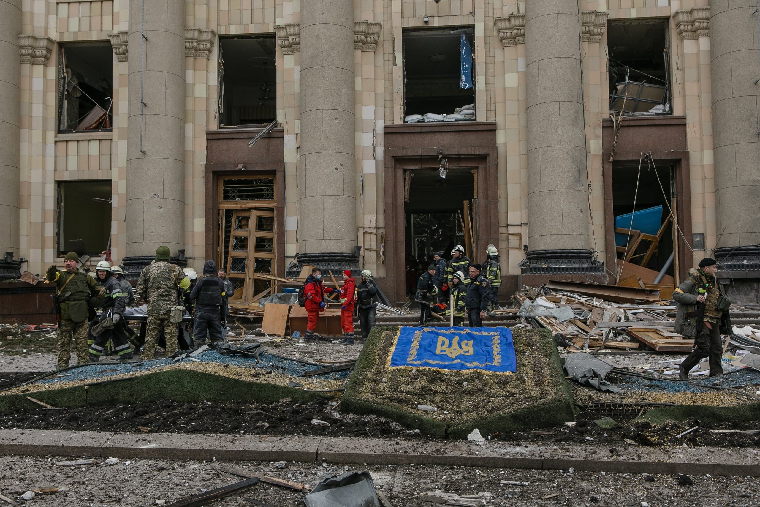 Разрушенное здание с колоннами, выбитые окна и двери, обломки на земле. Спасатели, медики и военные осматривают место. На переднем плане лежит украинский флаг с гербом.