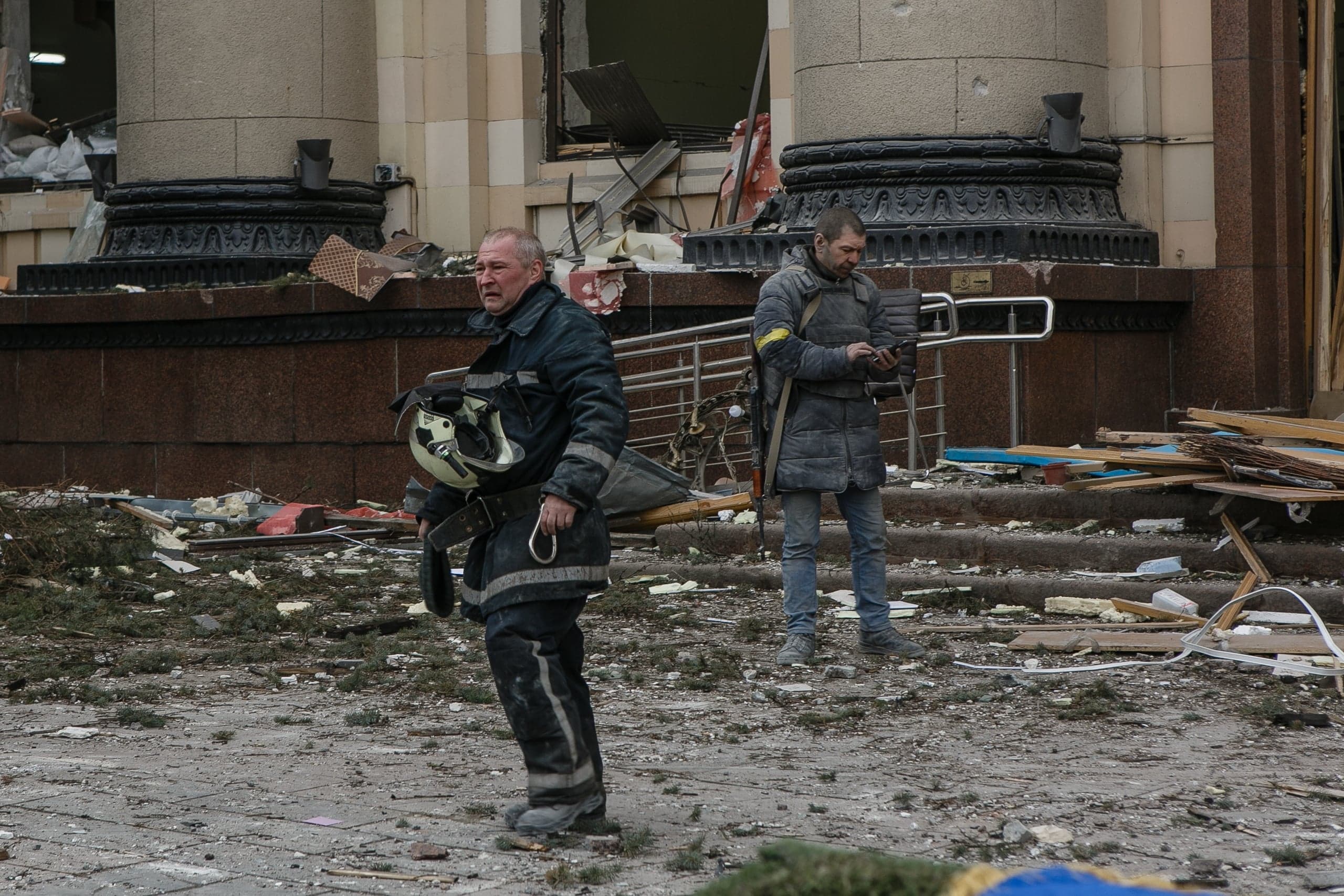 Пожарный с каской и вооруженный мужчина среди обломков перед поврежденным зданием.