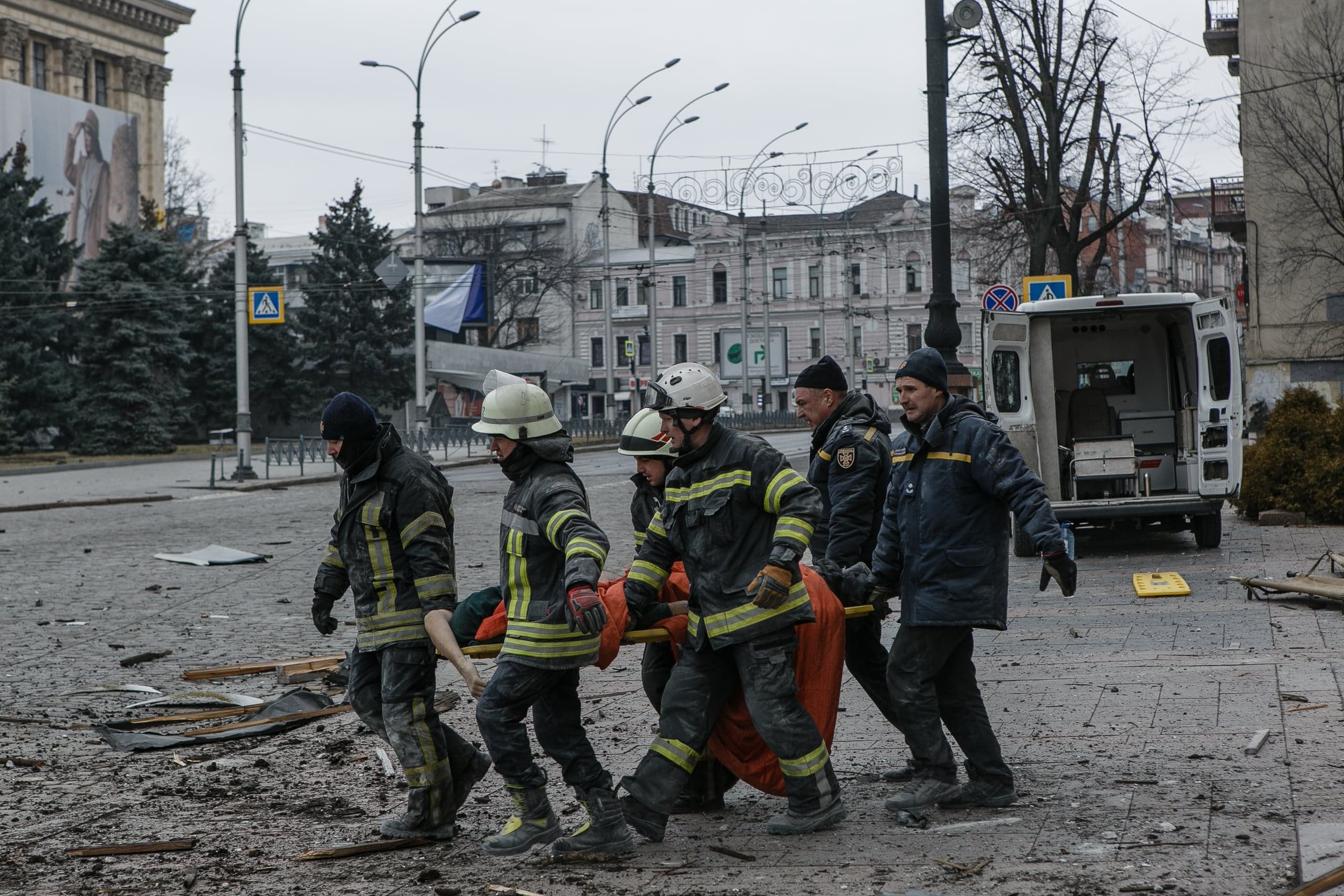 Шесть спасателей несут пострадавшего на носилках через улицу, покрытую обломками. На заднем плане припаркована машина скорой помощи.