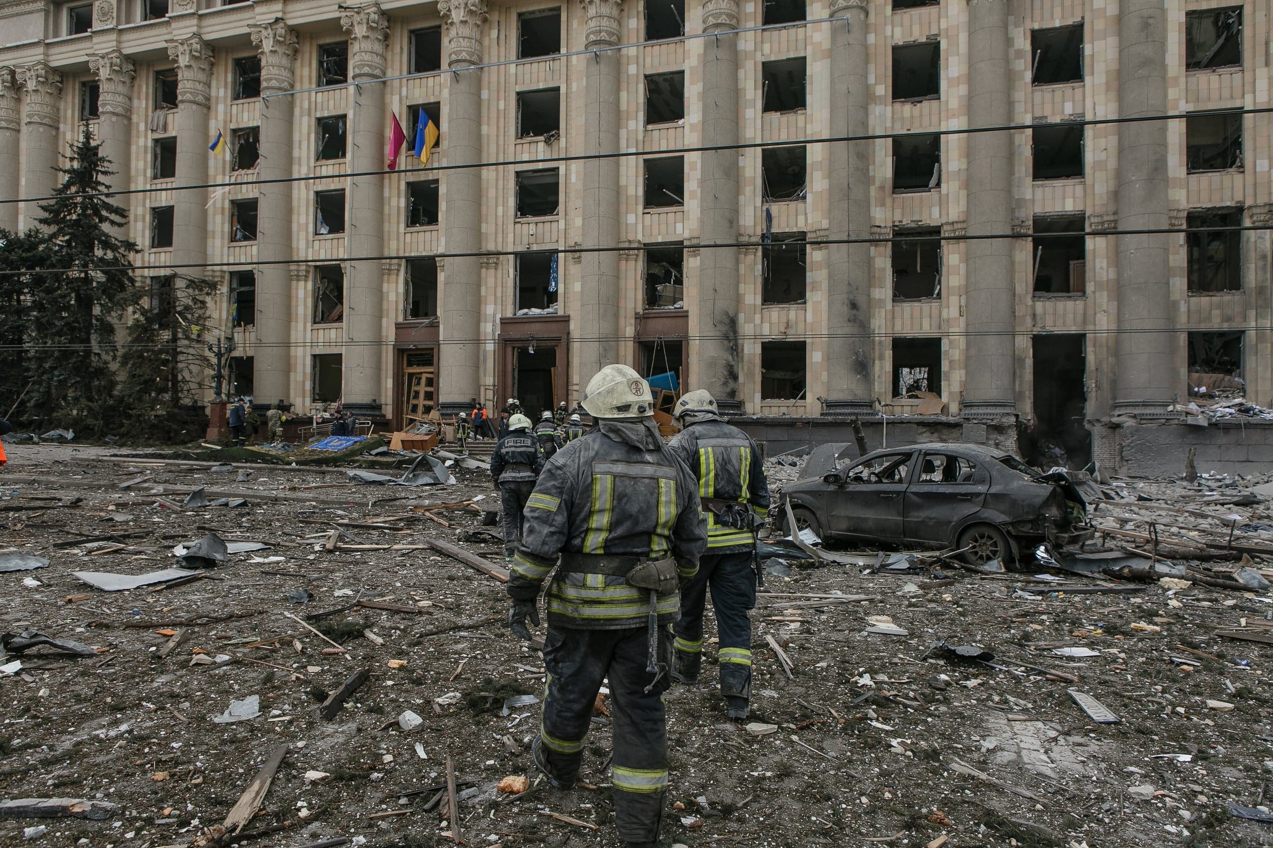 Двое спасателей идут по заваленному обломками двору перед сильно поврежденным зданием с выбитыми окнами. Рядом стоит разбитый автомобиль.