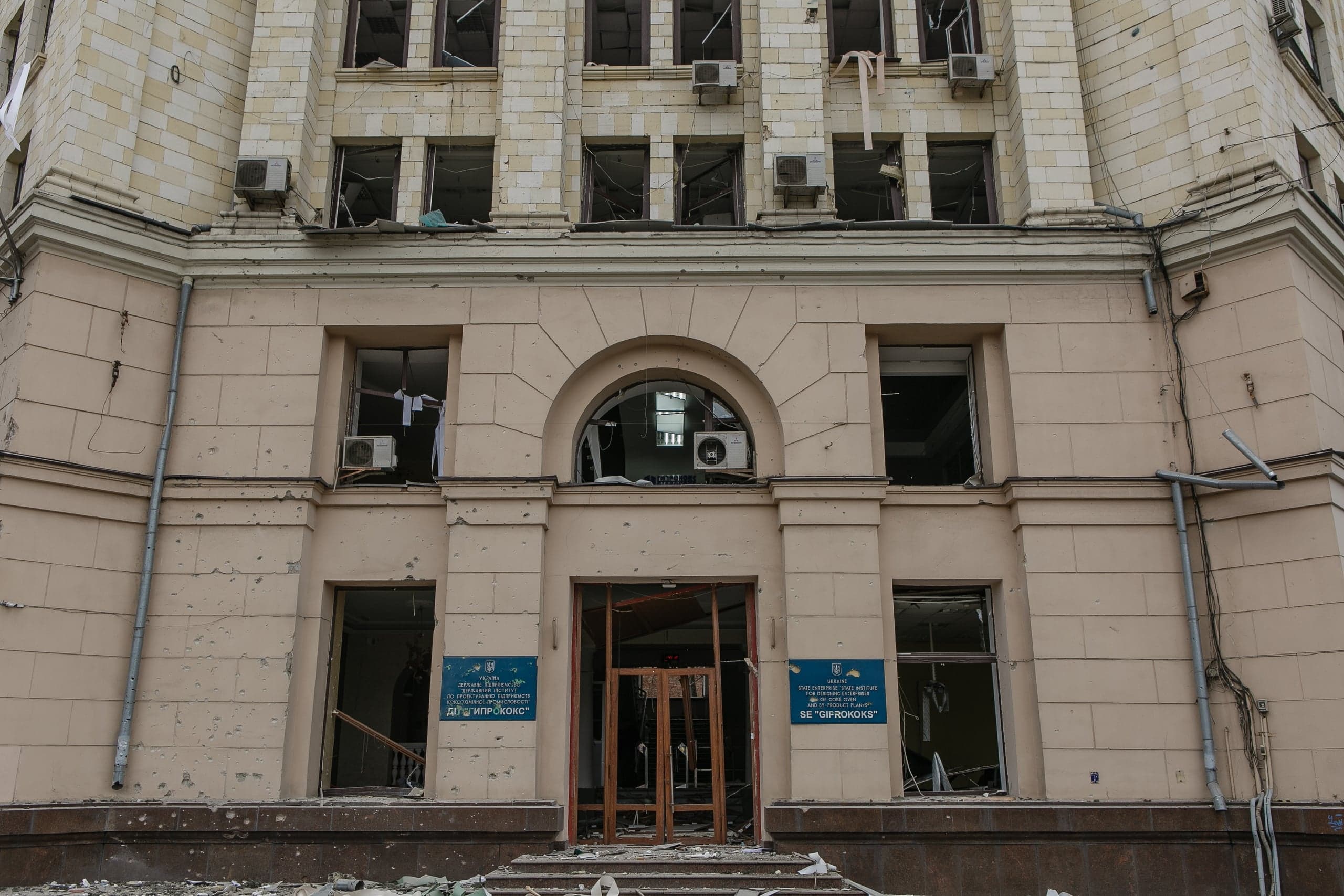 Сильно поврежденный фасад здания: разбитые окна, выбитая входная дверь, многочисленные щербины от осколков на стенах. Возле входа таблички "ДП "ГІПРОКОКС"" и "SE "GIPROKOKS"".