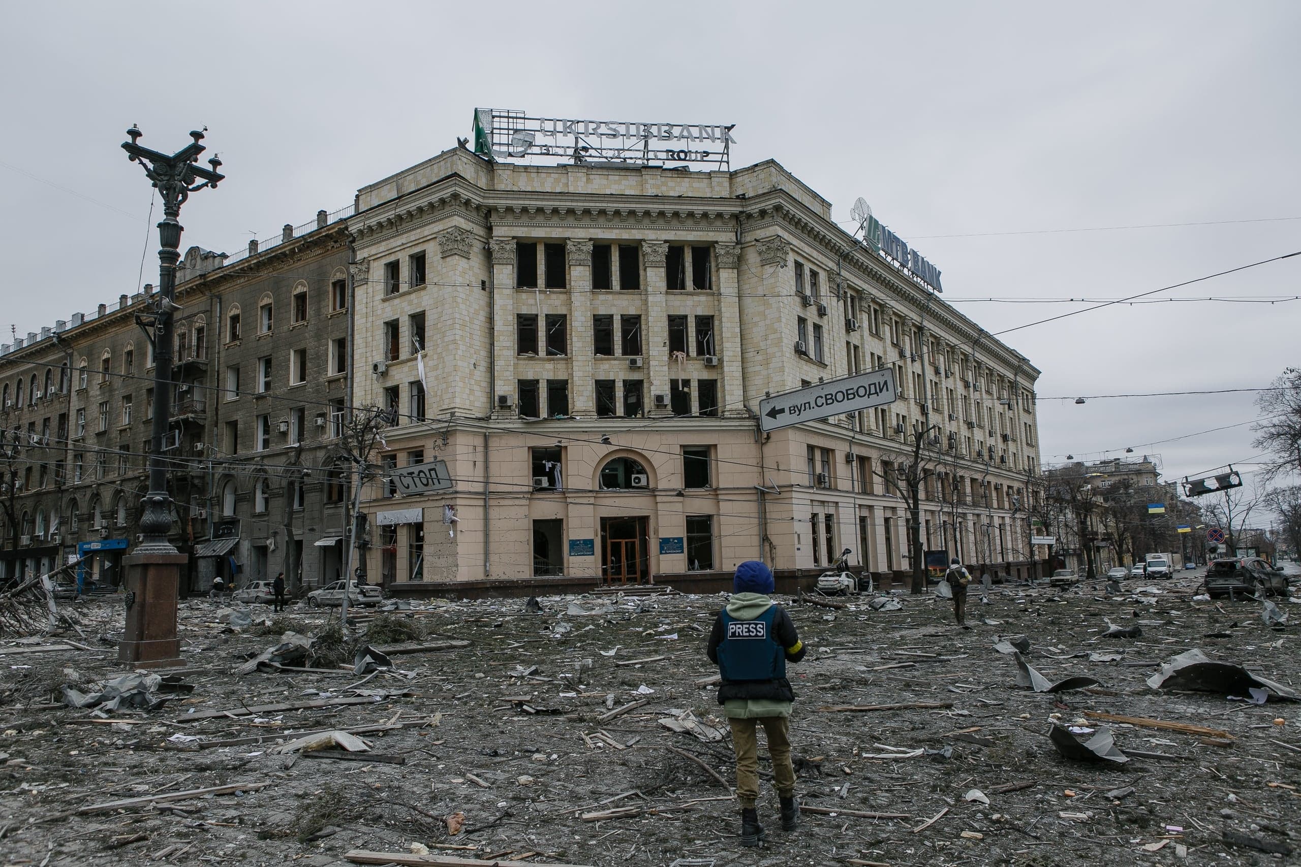 Журналист в жилете "PRESS" стоит среди обломков перед сильно поврежденным зданием. На здании вывеска "FIRST UKR. BANK GROUP" и указатель "вул. Свободи".