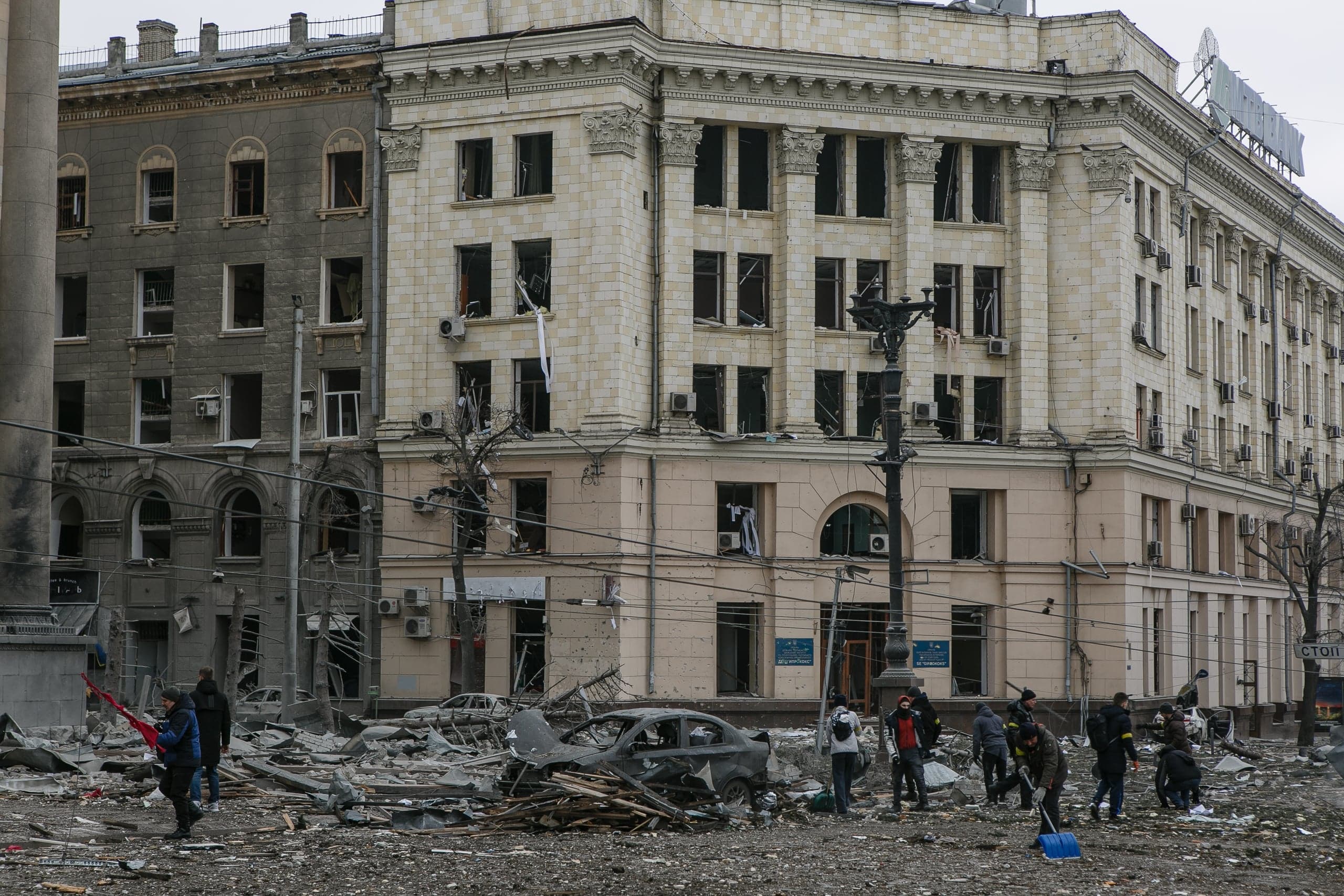 Разрушенные здания с выбитыми окнами, на переднем плане обломки, сгоревшая машина и группа людей, расчищающих завалы.