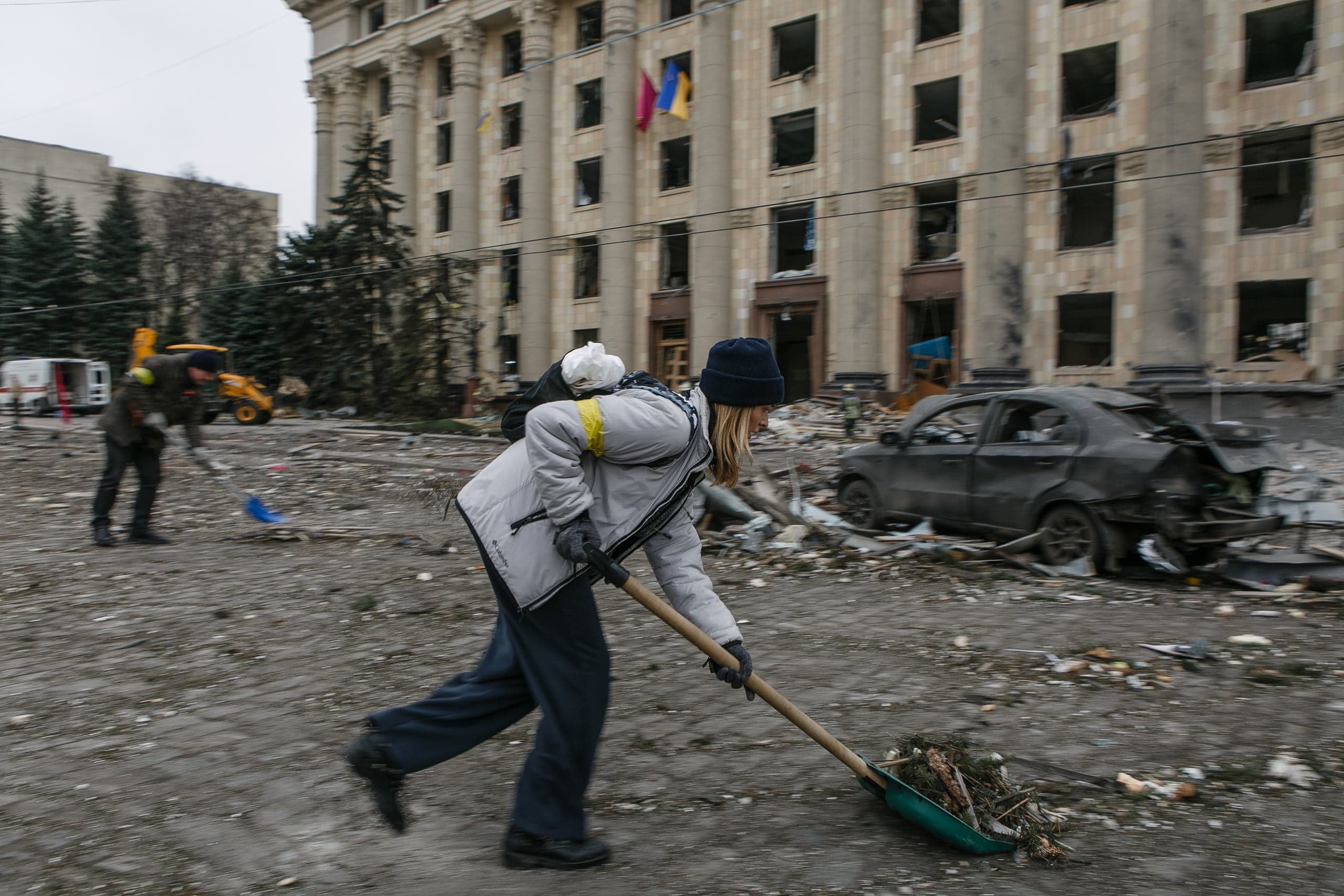 Волонтер убирает обломки лопатой на площади перед сильно поврежденным зданием с флагами Украины. Справа стоит сгоревший автомобиль.