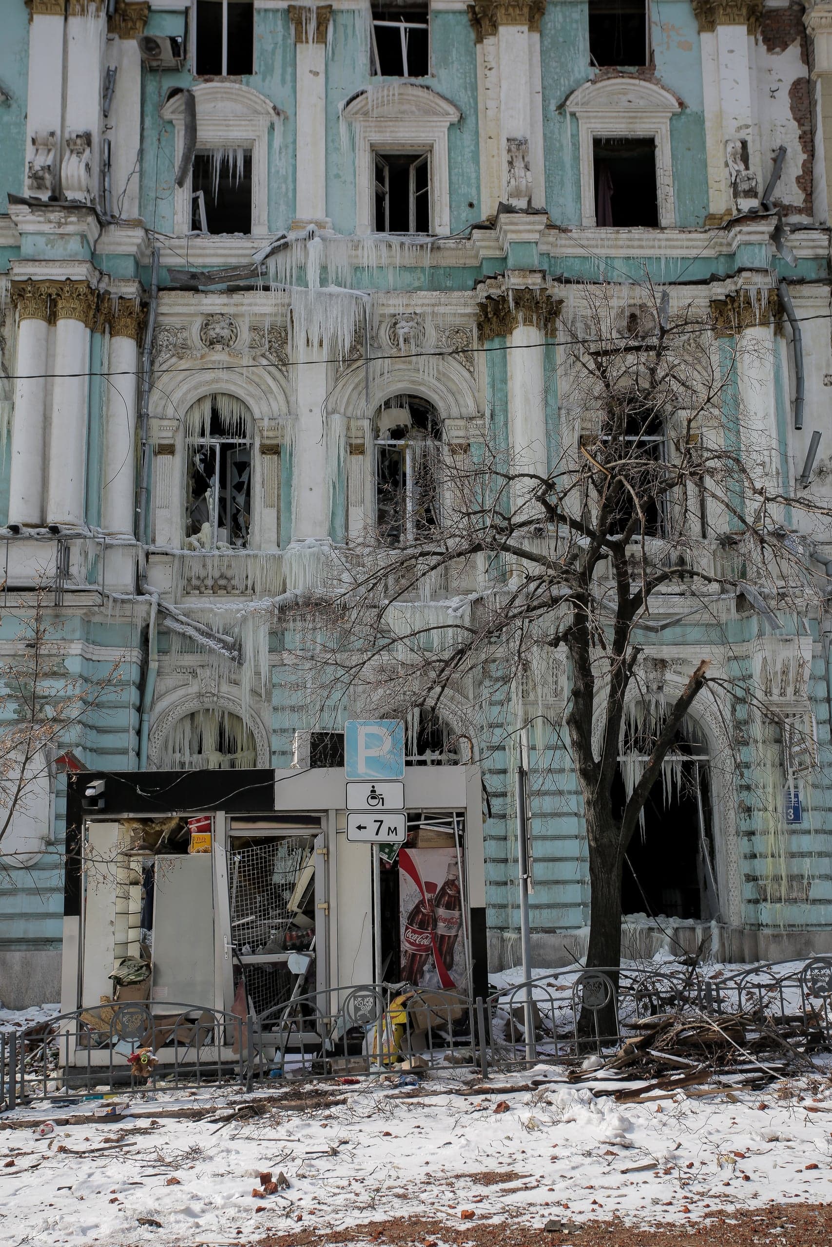 Разрушенный фасад старинного здания с разбитыми окнами, покрытый длинными сосульками. На переднем плане голое дерево, снег и разгромленный уличный киоск. Знак парковки "P", "← 7 M".