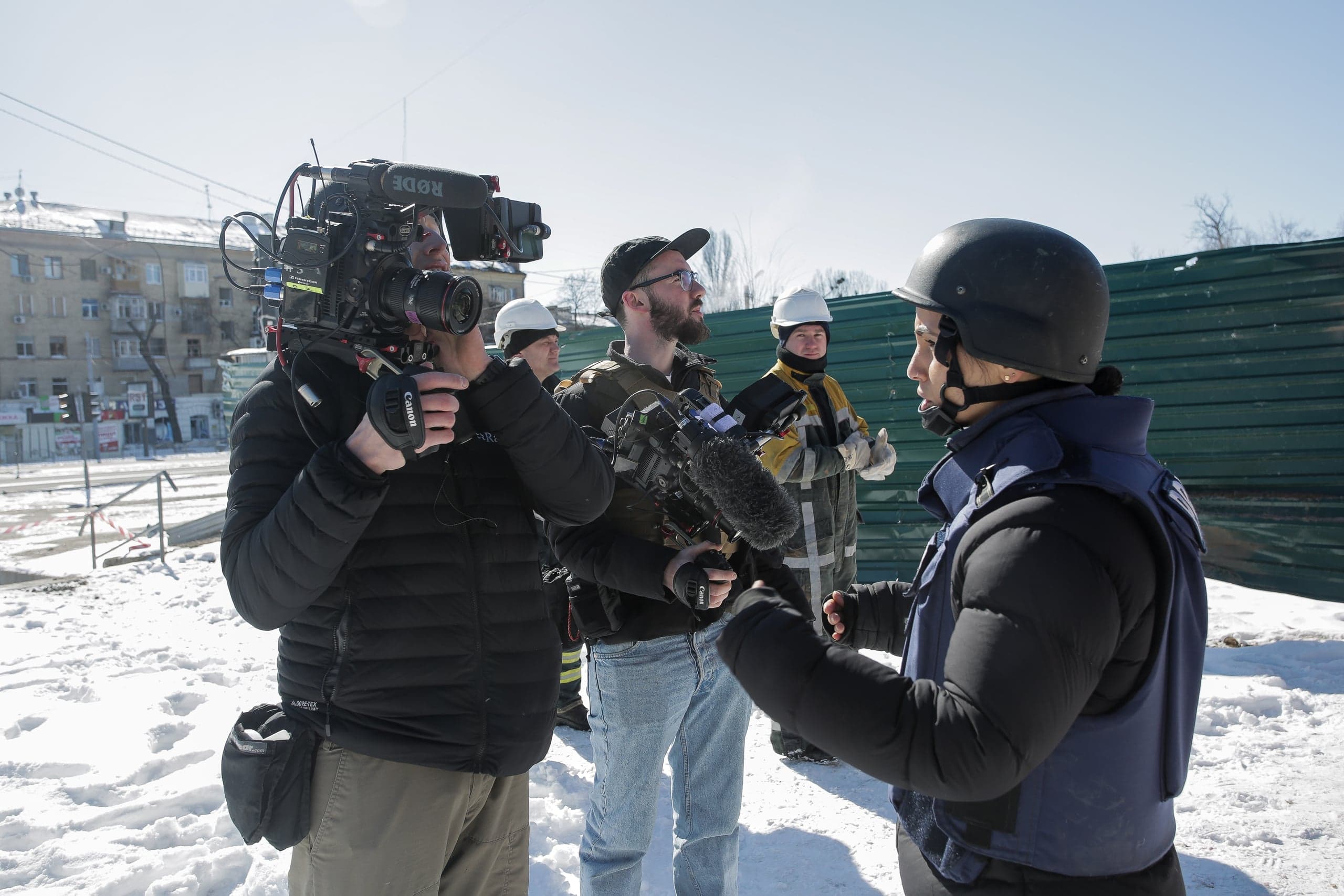 Группа из 5 людей стоит на снегу, один человек снимает профессиональной камерой другого человека в каске и синем жилете