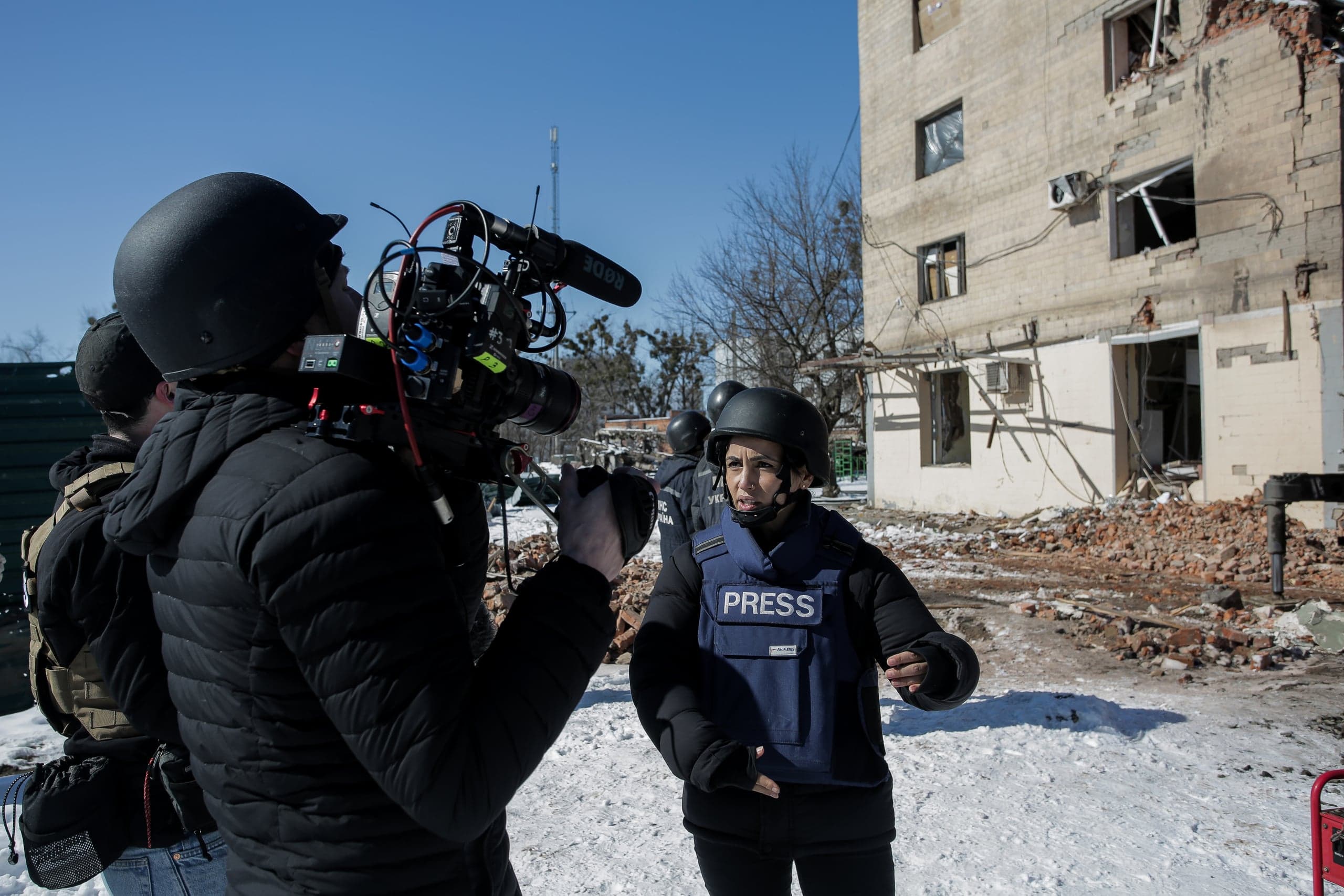 Журналист в бронежилете с надписью "PRESS" и оператор в шлемах снимают репортаж на фоне разрушенного здания.
