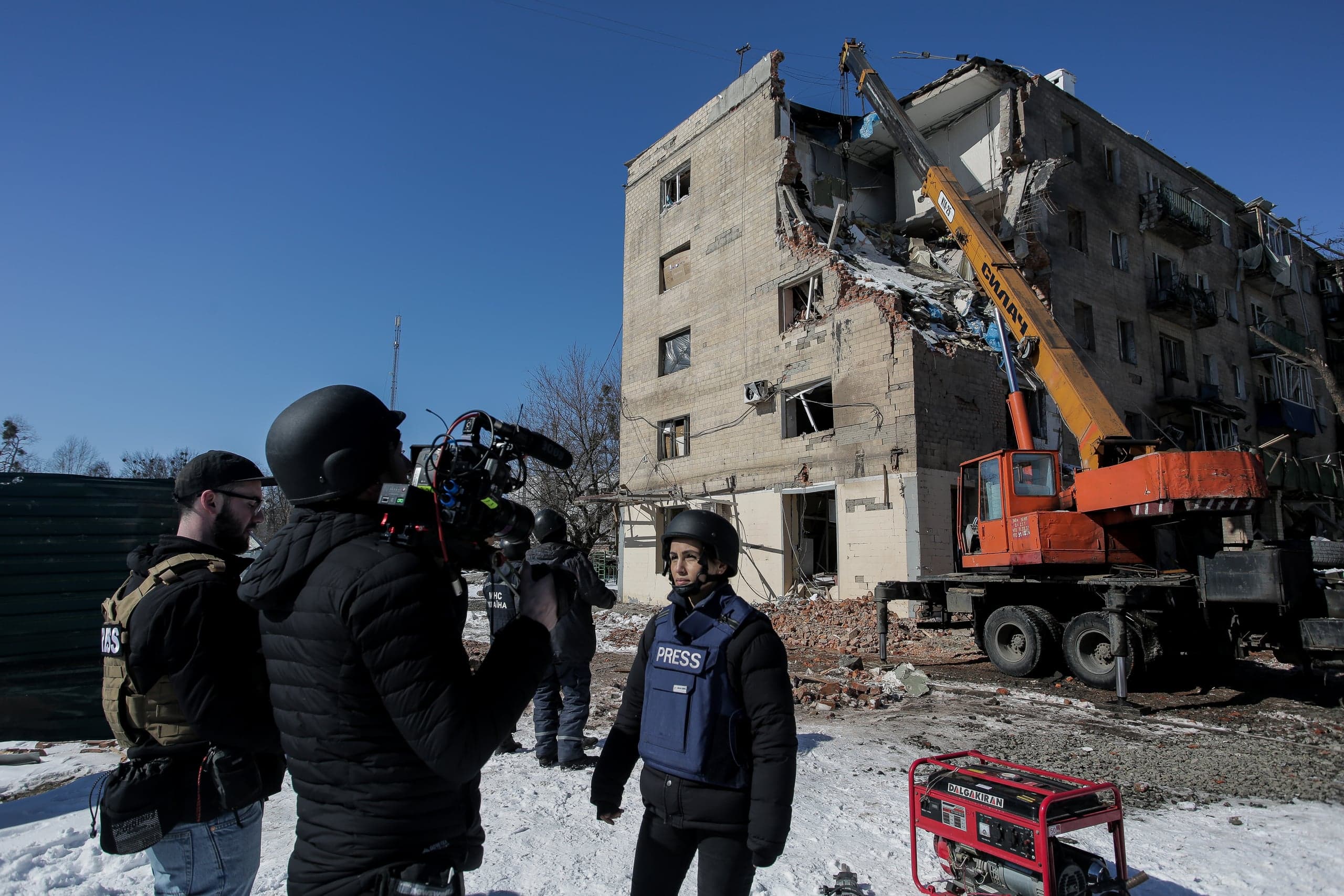 Репортеры снимают разрушенный жилой дом. Журналистка в жилете "PRESS" стоит перед оператором. Рядом с обломками работает строительный кран.