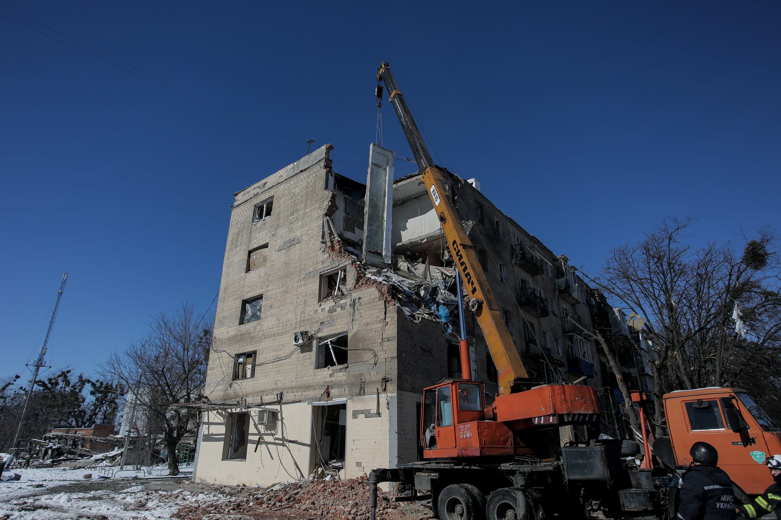 Кран демонтирует секцию разрушенного многоквартирного дома под чистым голубым небом. На переднем плане спасатели ГСЧС.