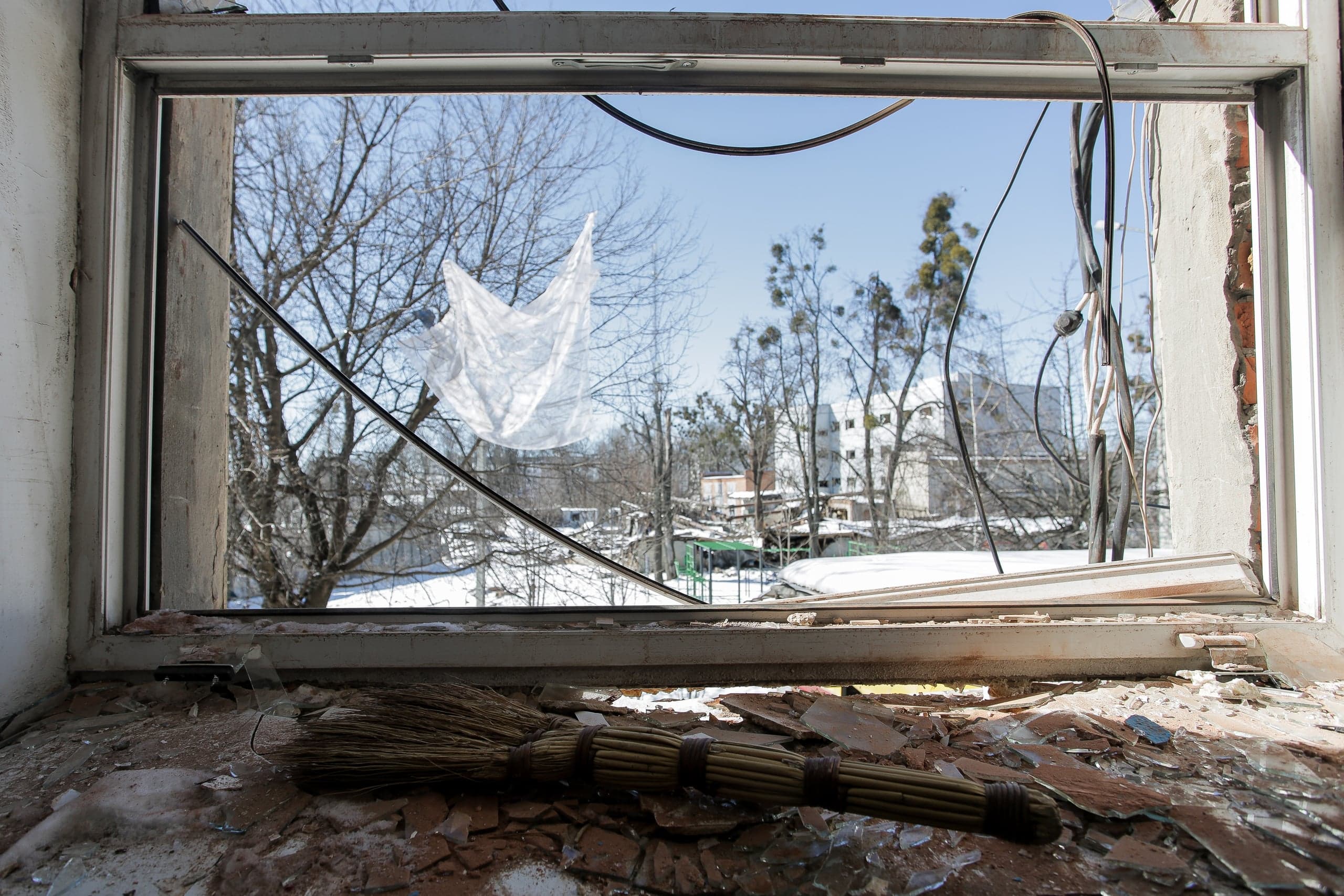 Разбитое оконное отверстие; подоконник завален осколками стекла и веником. Сквозь проем виден зимний городской пейзаж с деревьями и белыми зданиями.