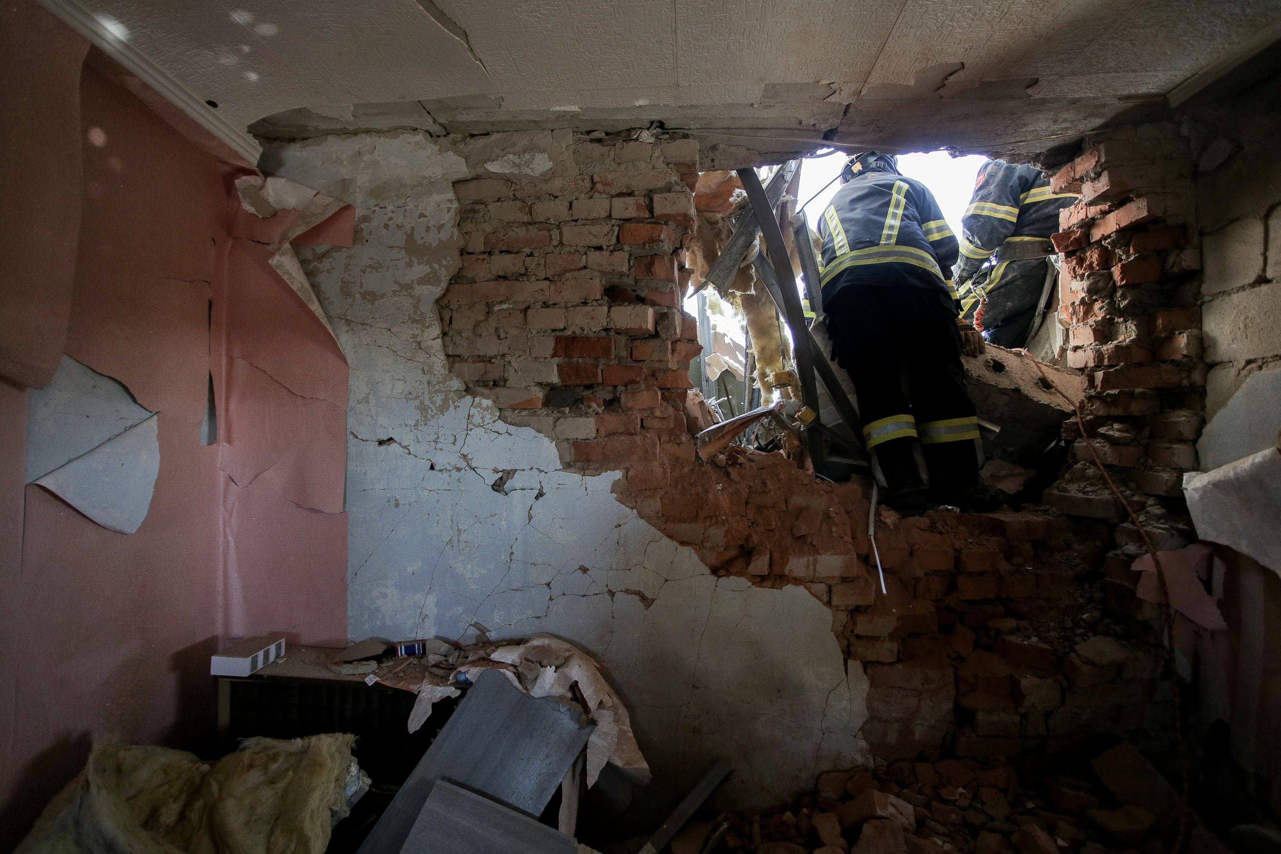 Спасатели пробираются через пролом в разрушенной кирпичной стене здания.