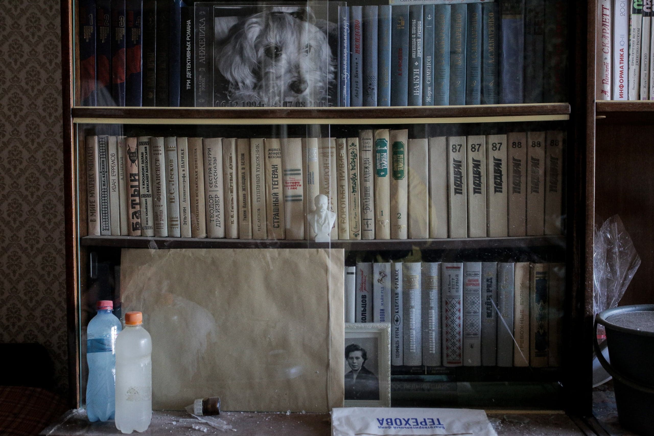 Застекленный книжный шкаф с рядами старых, пыльных книг. На полках: обложка с изображением собаки, бюст, две пластиковые бутылки, фотография и надпись "ТЕРЕХОВА".