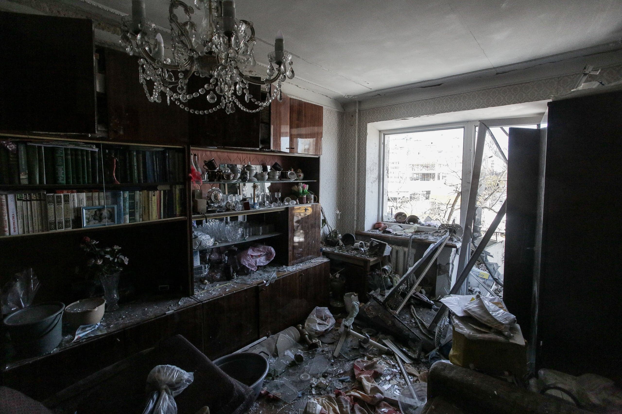 Разрушенная комната, на потолке стеклянная люстра, вдоль стены большой шкаф с книгами и посудой, большое окно разбито