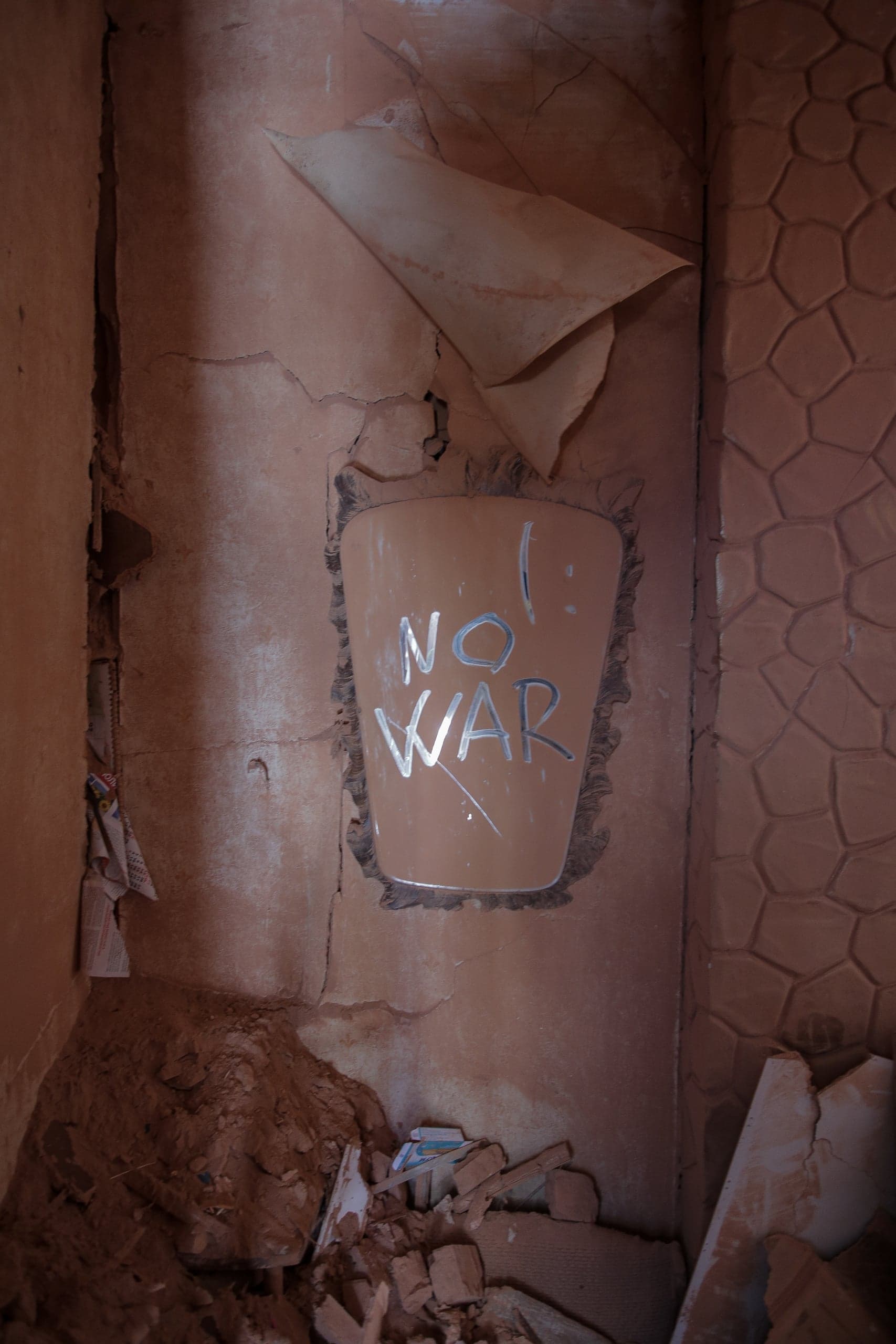 Разрушенная глиняная стена с облупившейся штукатуркой и надписью "NO WAR". На полу куча обломков.