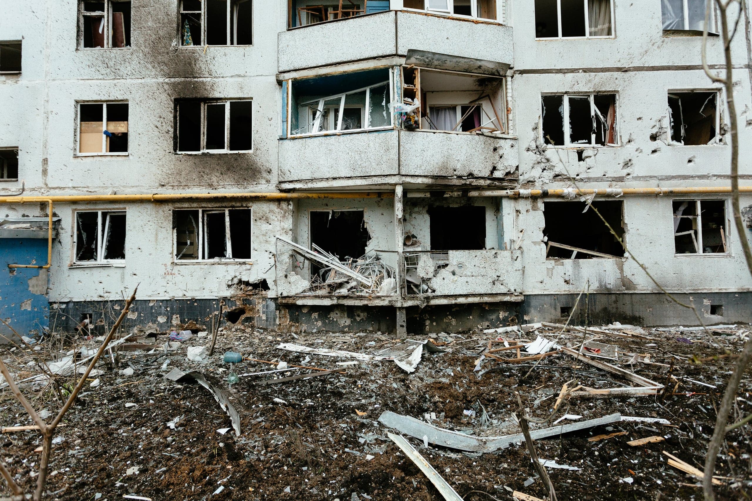 Поврежденный обстрелом фасад жилого дома: выбиты окна, разрушены балконы, стены покрыты следами взрывов, перед домом груды обломков.