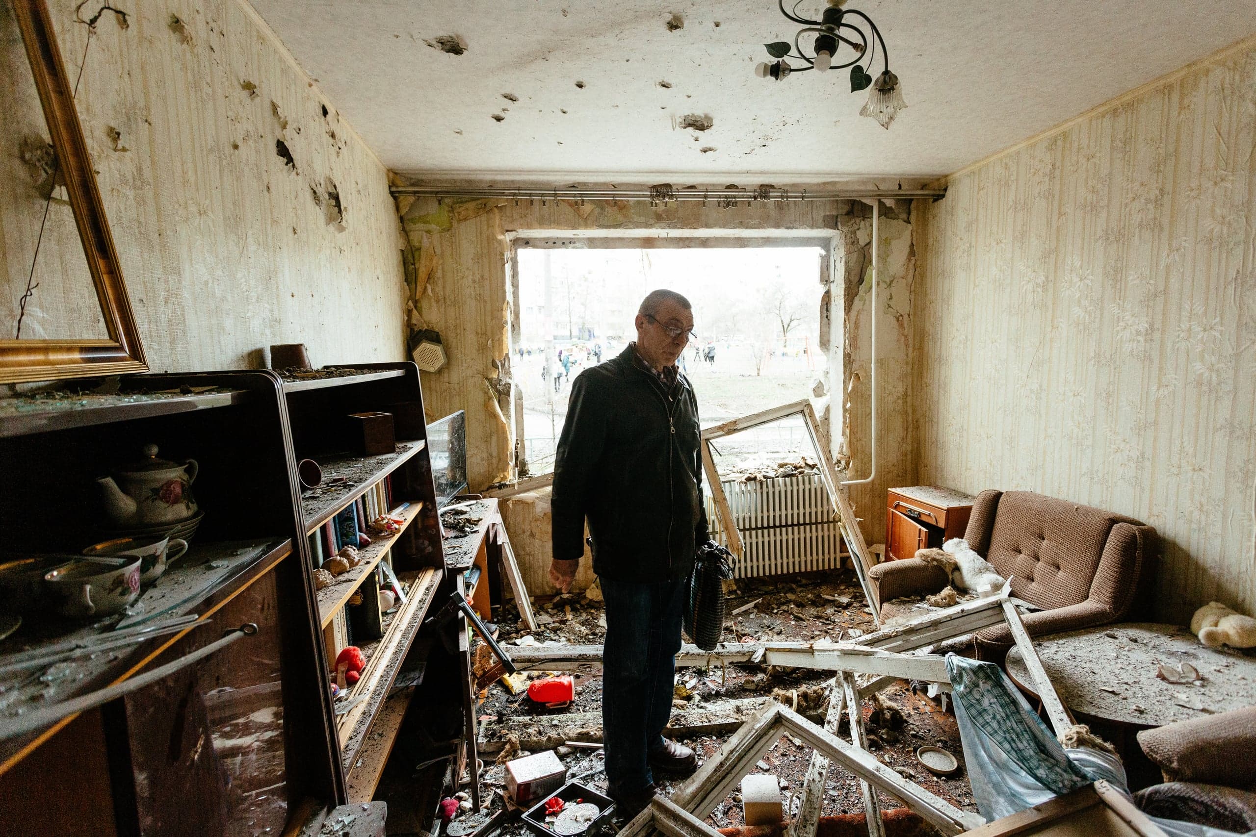 Мужчина стоит в сильно разрушенной жилой комнате: выбитое окно, пробоины в стенах, пол завален обломками и мебелью.