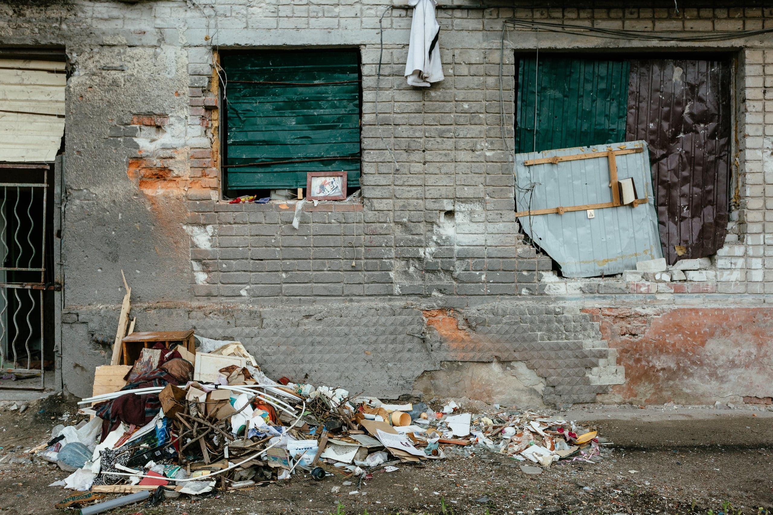 Ветхая кирпичная стена с заколоченными окнами; перед стеной — большая куча мусора и хлама.