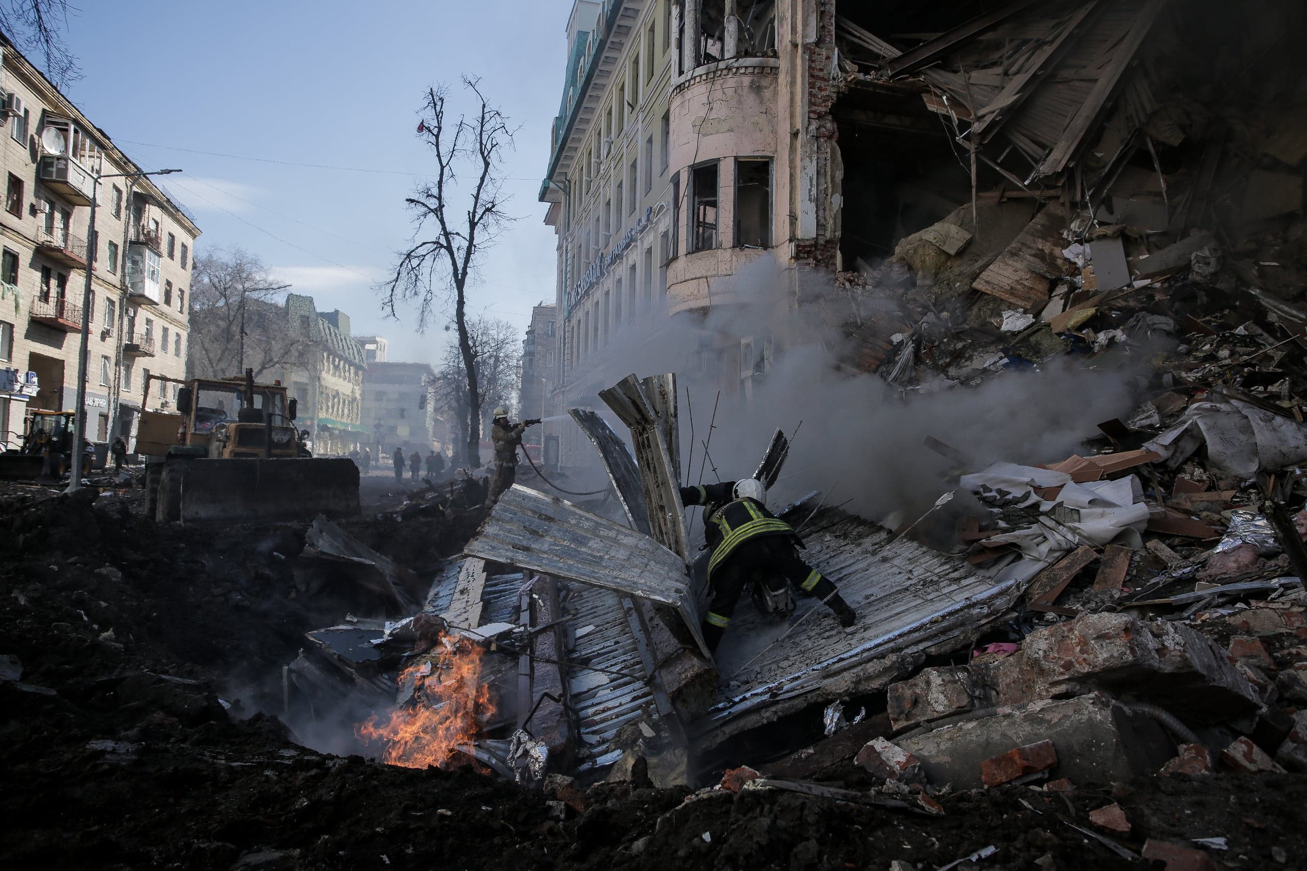 Спасатели работают среди обломков разрушенного здания на городской улице, видно пламя и дым, на заднем плане бульдозер.