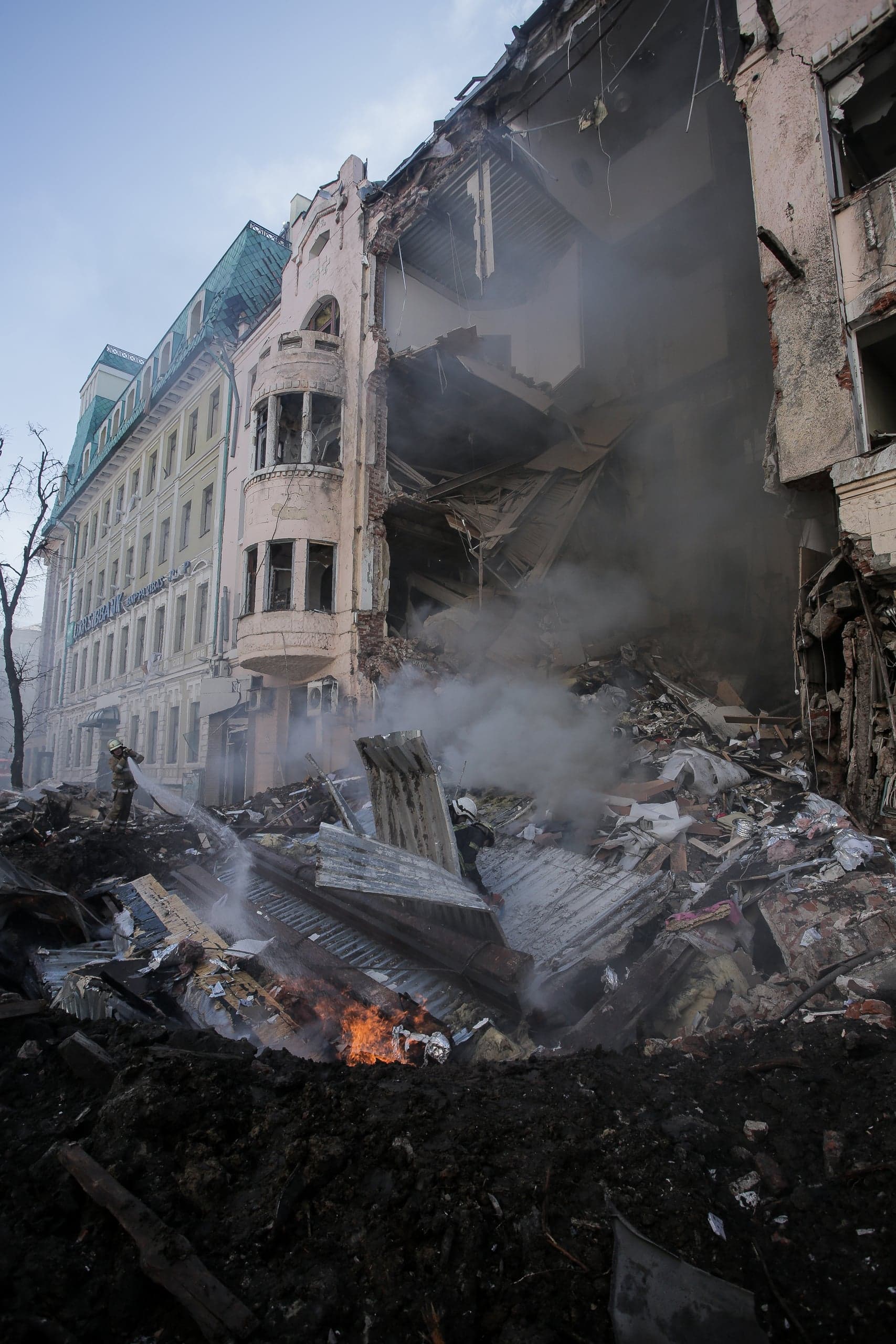 Разрушенное здание с обвалившейся секцией, обломки на переднем плане, дым и открытое пламя. Пожарный тушит очаг возгорания.