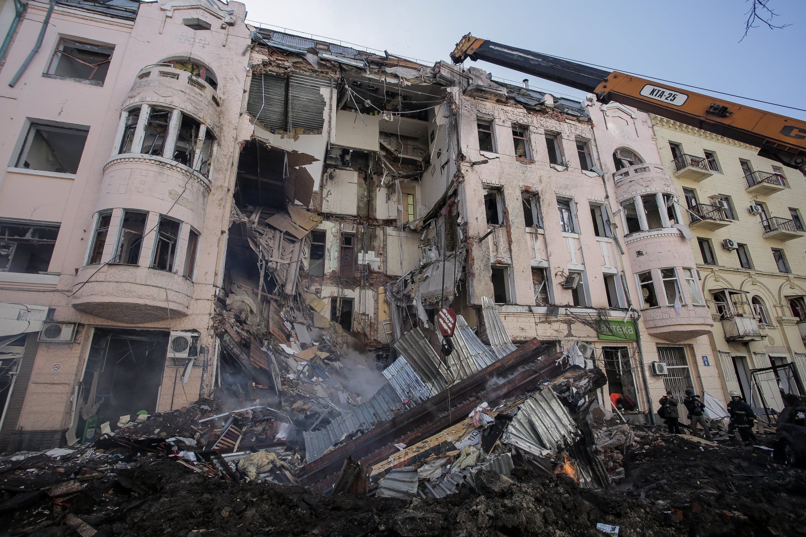 Сильно поврежденный многоэтажный жилой дом с обрушенной центральной секцией. Над грудой обломков работает стрела крана "КТА-25", внизу видны спасатели.