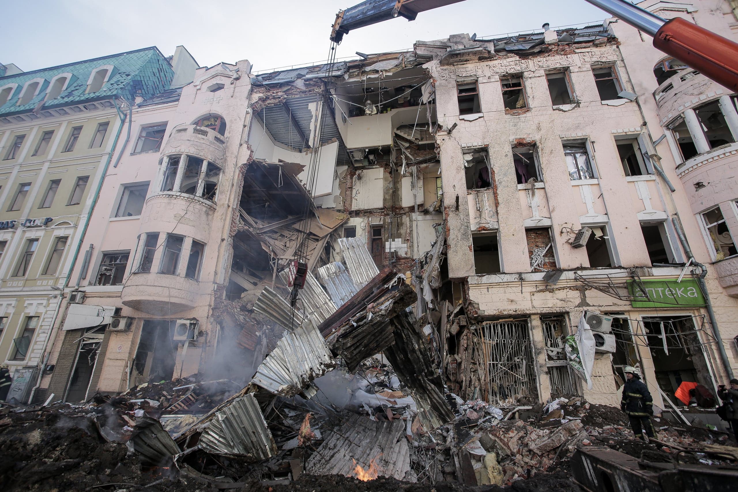 Разрушенное здание с обвалившейся центральной частью, горы обломков и дым. Над завалами работает стрела крана, внизу спасатели. Видна вывеска "АПТЕКА".