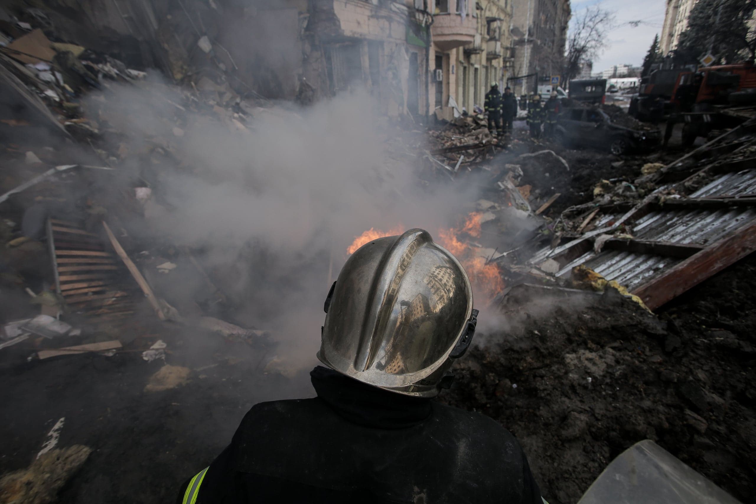 Пожарный в шлеме стоит среди дыма, огня и обломков разрушенной улицы. На заднем плане видны другие спасатели.