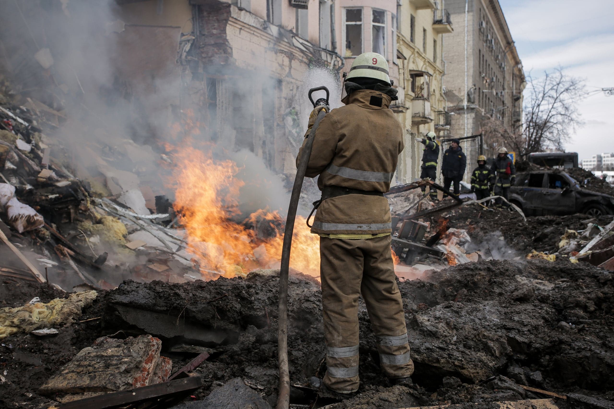 Пожарный тушит сильный огонь на развалинах разрушенного здания; вокруг дым, груды обломков и другой персонал.