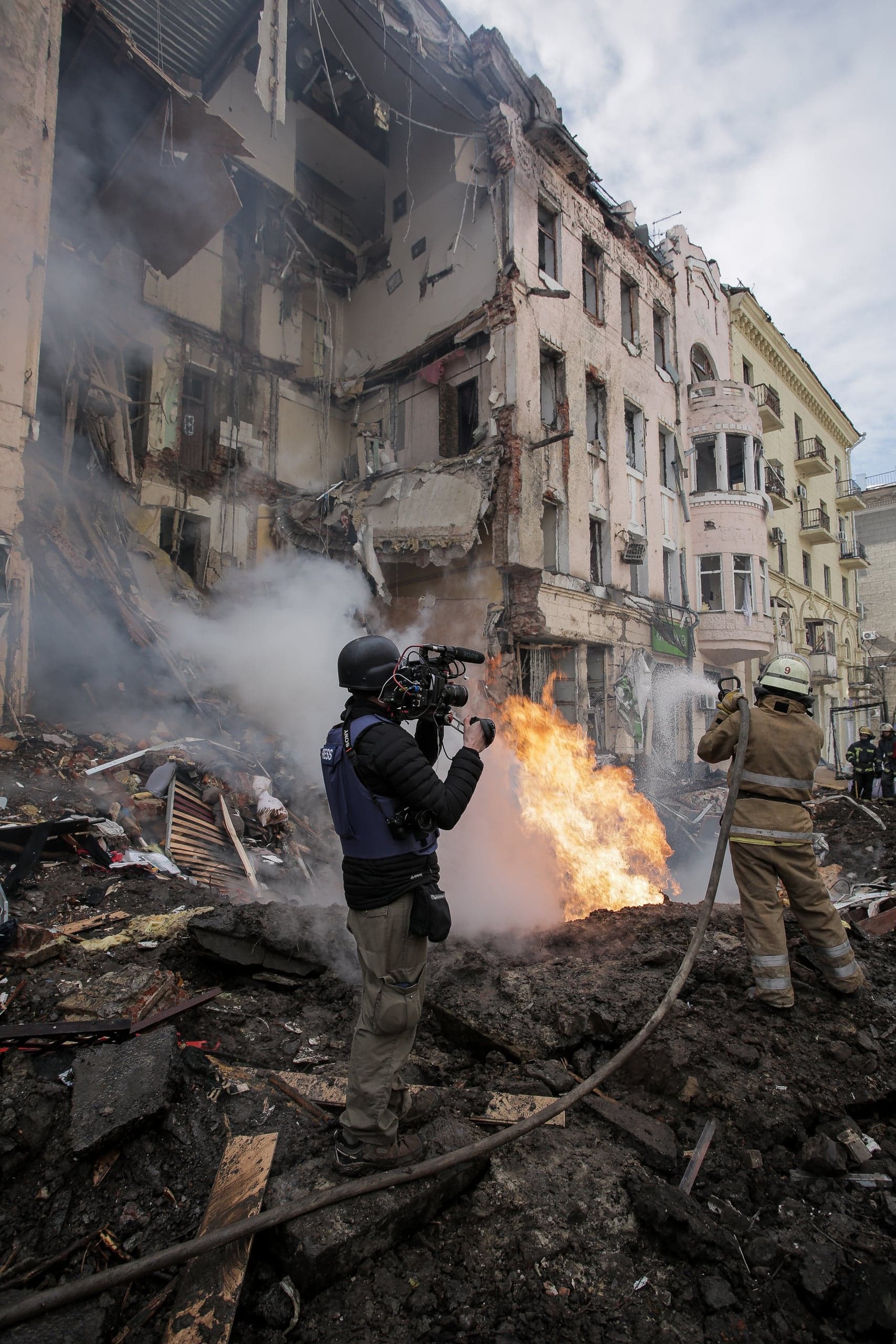 Журналист снимает сильный пожар на фоне разрушенного здания; пожарный тушит пламя водой.