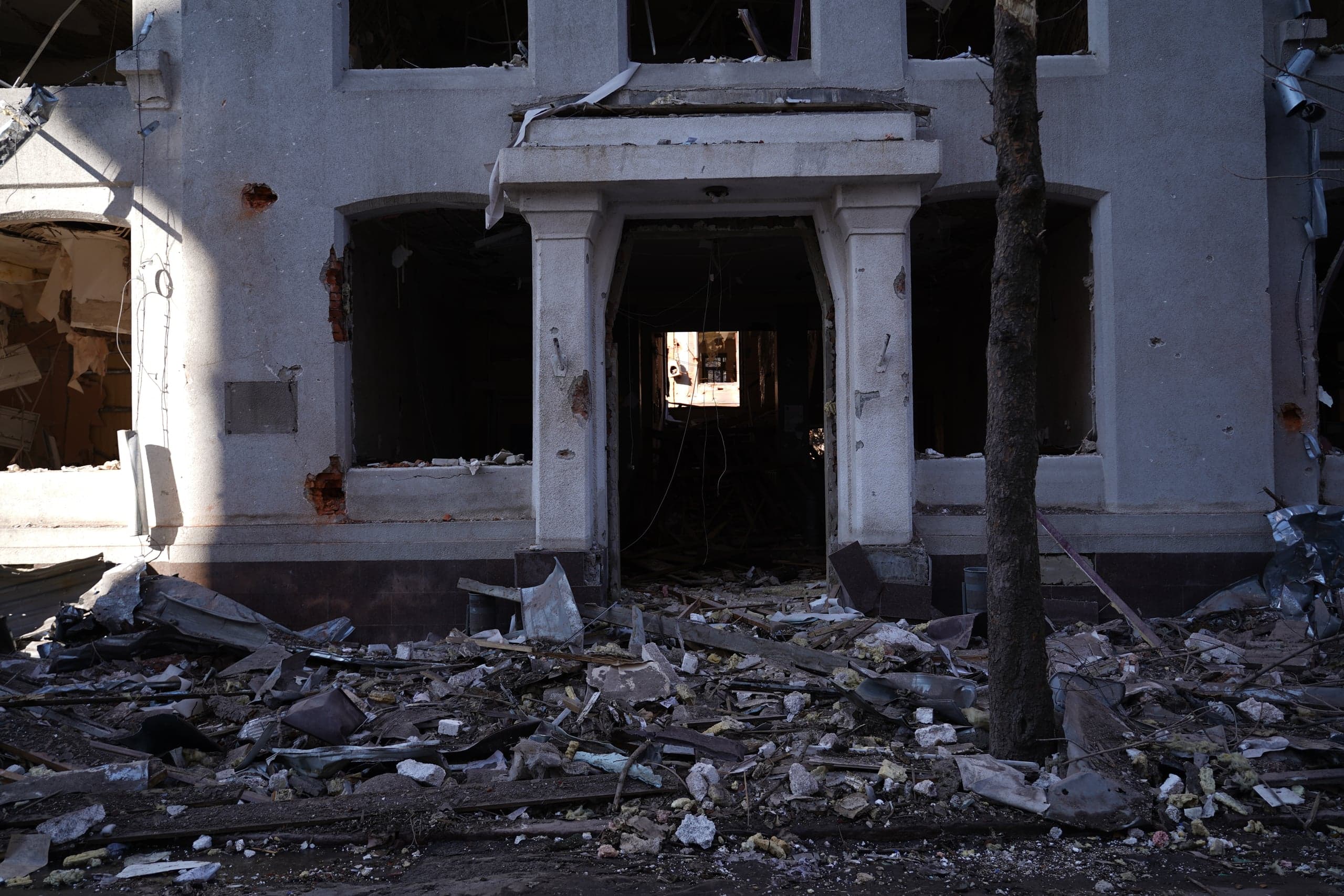 Фасад разрушенного серого здания с колоннами и пустыми оконными проемами. Перед входом — огромная гора строительного мусора и обломков. Левая часть стены ярко освещена солнцем.