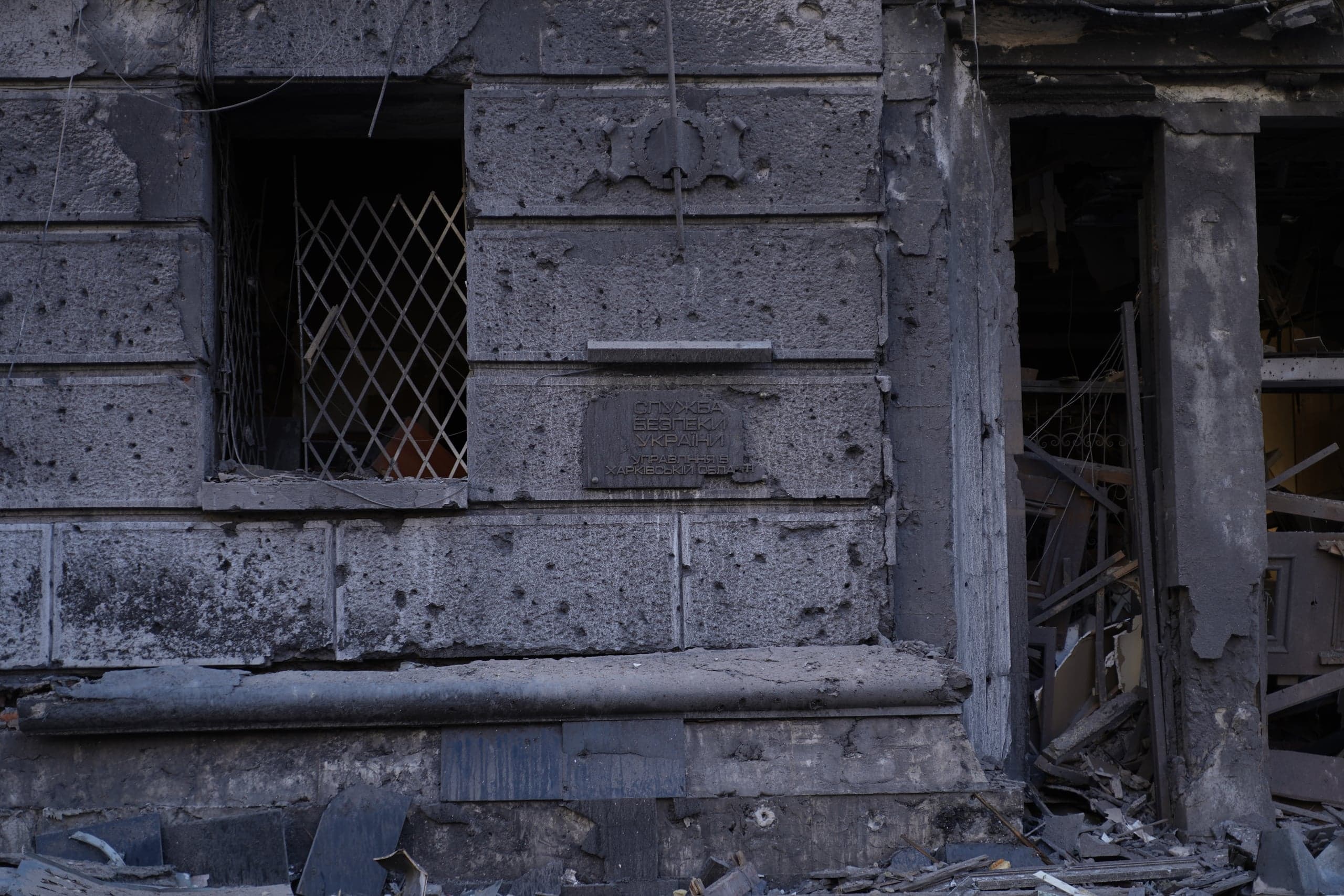 Обгоревший и иссеченный осколками фасад здания Управления СБУ в Харьковской области. На стене видна табличка с надписью «Служба безпеки України» и зарешеченное окно. В правой части виден разрушенный дверной проем с грудой обломков.