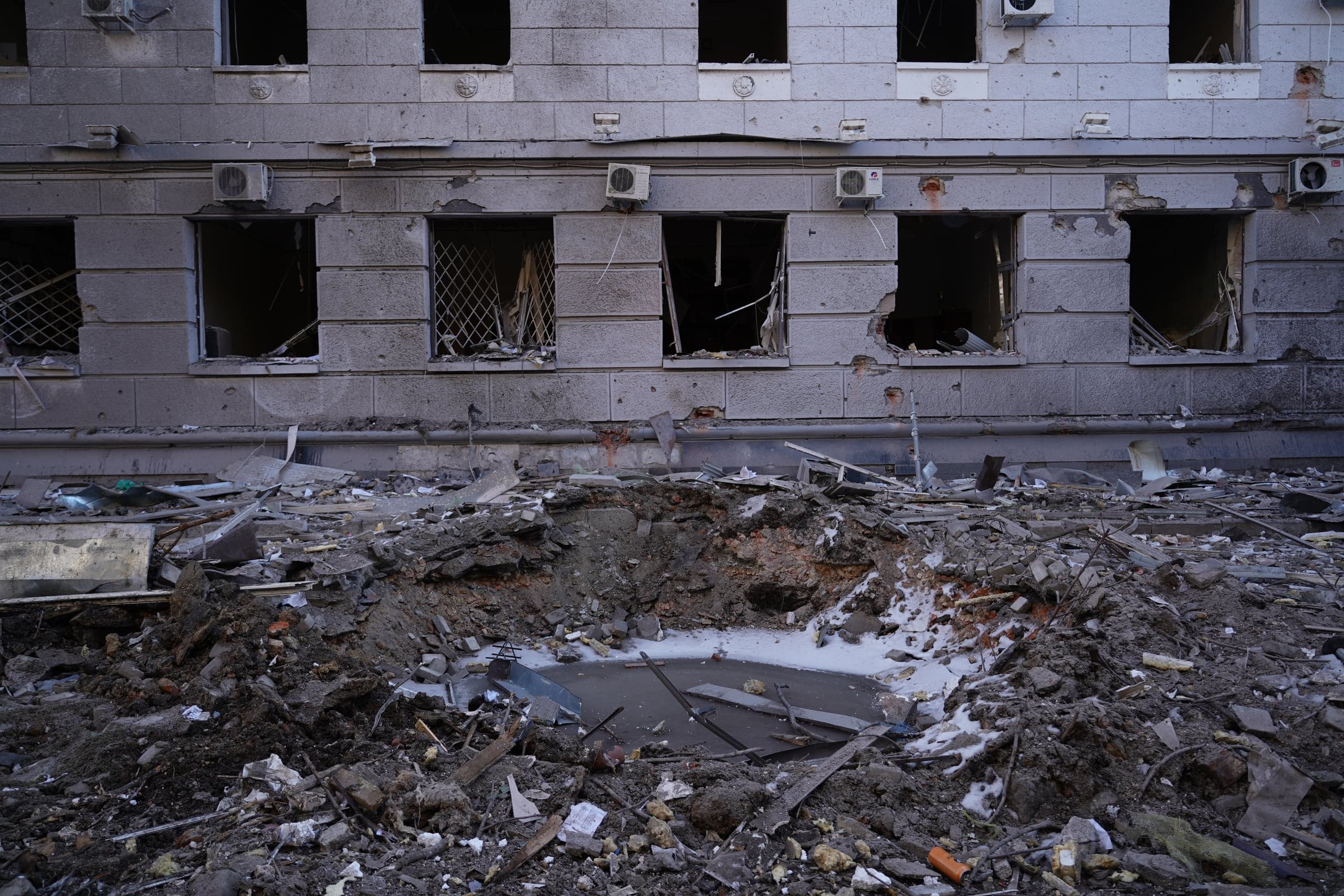 Большая воронка от взрыва, заполненная обломками и грязью, на фоне поврежденного серого фасада здания с выбитыми окнами. На стенах видны следы осколков и висят разбитые кондиционеры.