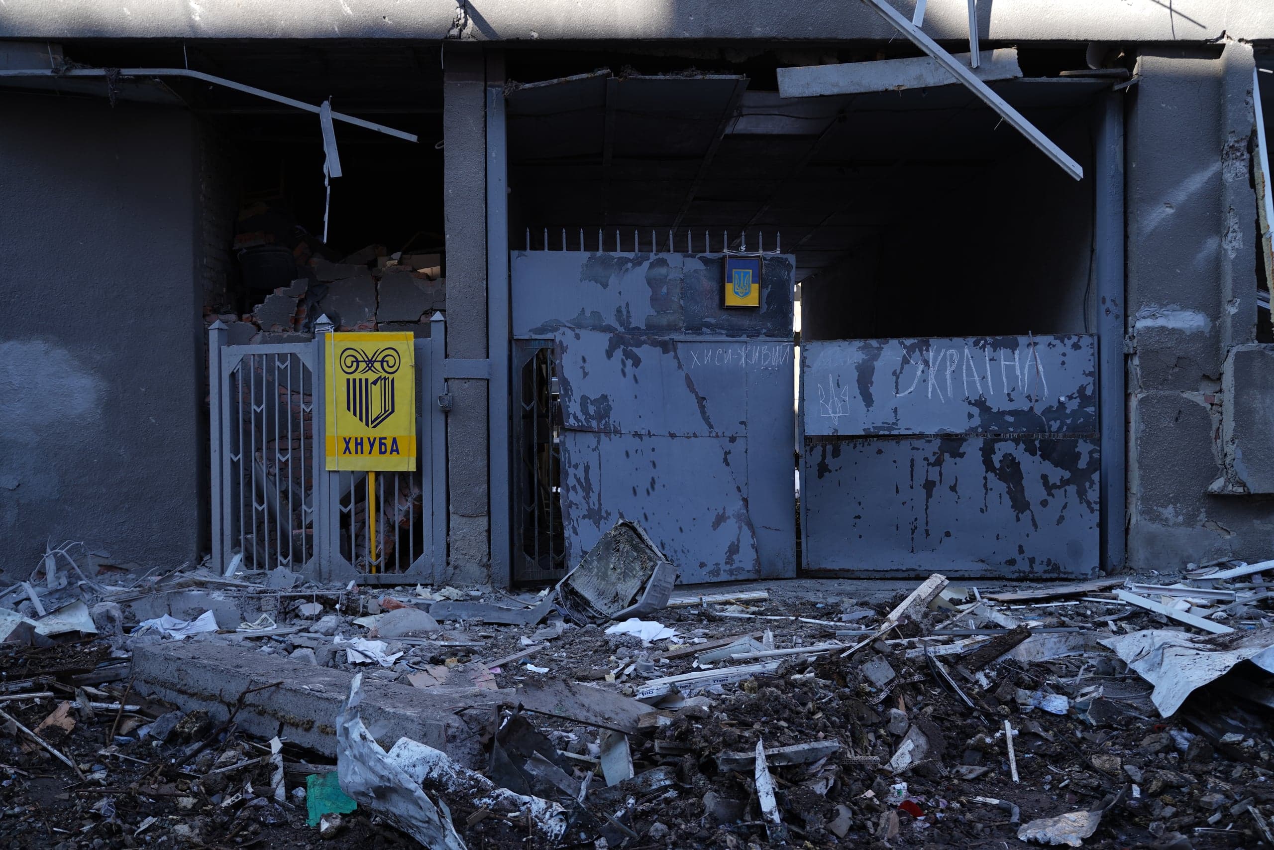 Вход в разрушенное здание ХНУБА (Харьковский национальный университет) в Харькове, заваленный обломками и мусором. На серых металлических воротах видна желтая табличка университета и граффити мелом, включая слово «УКРАИНА» и трезубец.